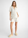 A woman models the Cozy Earth Women's Bamboo Stretch-Knit Short Sleeve Pajama Set, featuring a short-sleeved button-up shirt and shorts with a small multicolored design on a light background, paired with beige fuzzy slippers against a plain white backdrop. |Color:Sienna Tulips
