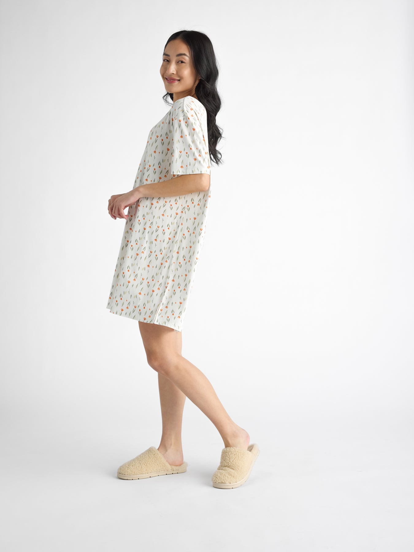 A woman in a Cozy Earth Women's Bamboo Stretch-Knit Sleep Dress and beige slippers stands sideways, smiling against a white background. 