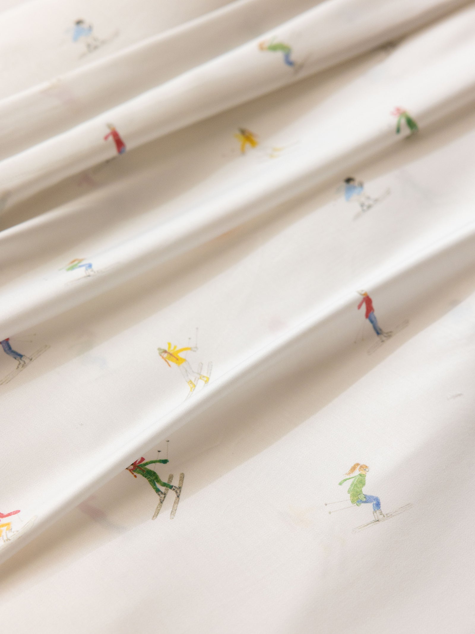 Close-up of the Bamboo Sheet Set by Cozy Earth, featuring a white fabric adorned with a whimsical pattern of small, colorful skiers in dynamic poses. The skiers sport outfits in red, yellow, green, and blue, adding a playful touch to the smooth material. 