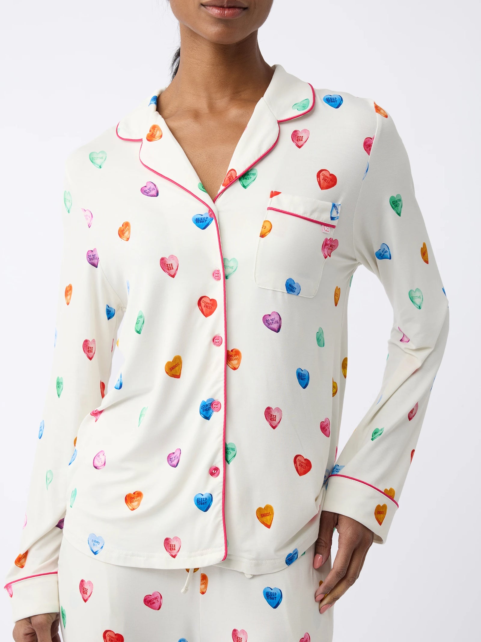 A person wears the Women's Long Sleeve Bamboo Pajama Top in Stretch-Knit by Cozy Earth, a white set adorned with colorful hearts and red piping on the collar, pocket, and cuffs. The background is plain white. 