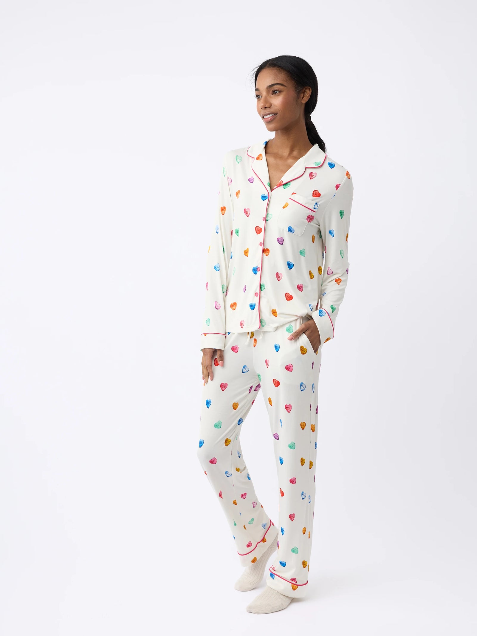 A person in a cream Cozy Earth Women's Stretch-Knit Bamboo Pajama Pant with multicolored hearts and red piping stands against a plain background, smiling with hands in pockets. 