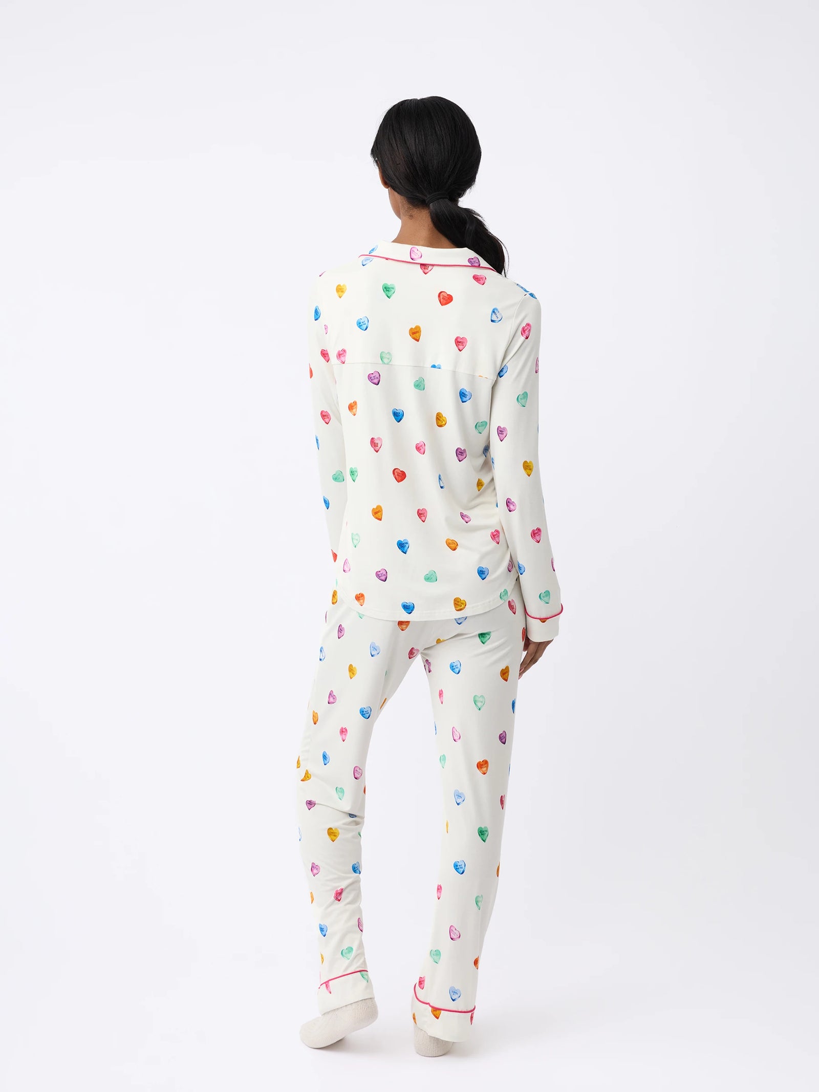 A woman stands facing away, wearing Cozy Earth's Women's Stretch-Knit Long Sleeve Bamboo Pajama Set in white with colorful heart patterns and red piping on the cuffs, set against a plain, light background. 