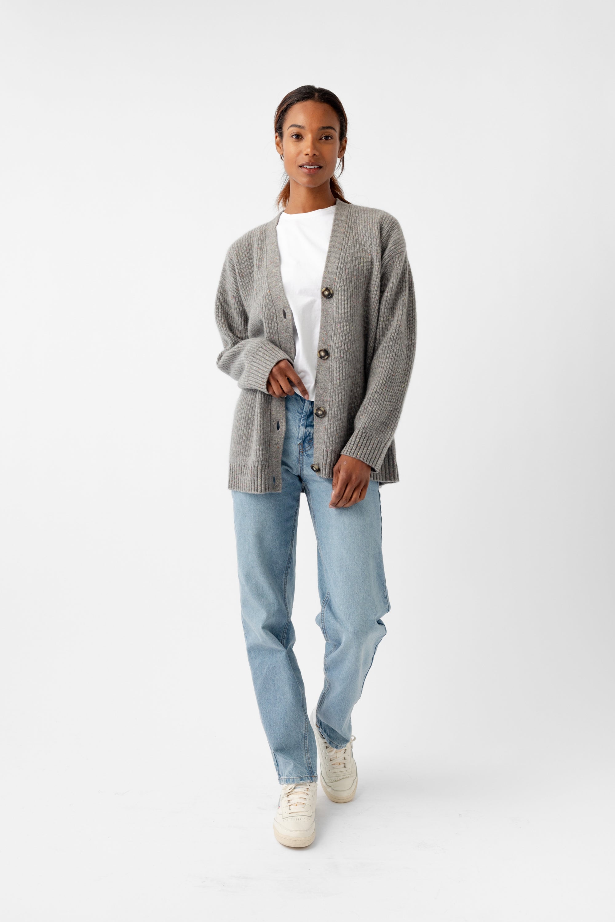 A person wears a Cozy Earth Oversized Sunday Cashmere Cardigan in gray over a white shirt, paired with light blue jeans and white sneakers. They are standing against a plain white background. 