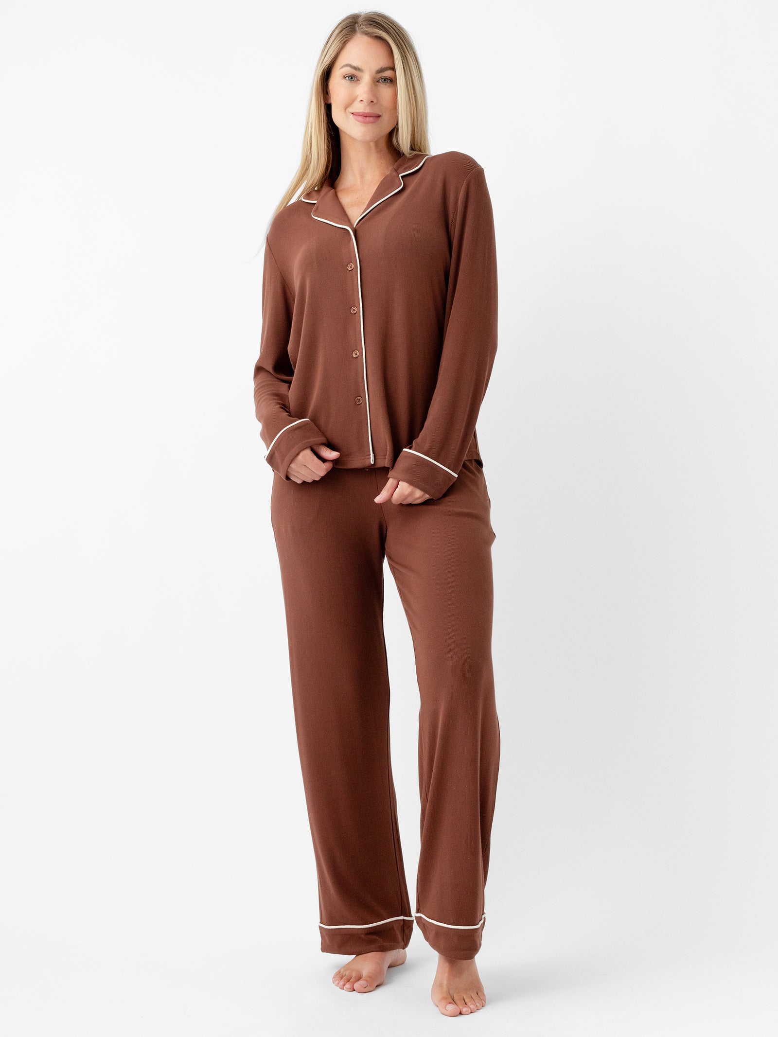 A woman stands barefoot against a plain white background wearing Cozy Earth’s Women's Bamboo Rib Knit Classic Pajama Pant in brown, featuring long sleeves and white piping. She has light-colored hair and is smiling slightly with her arms relaxed by her sides. 