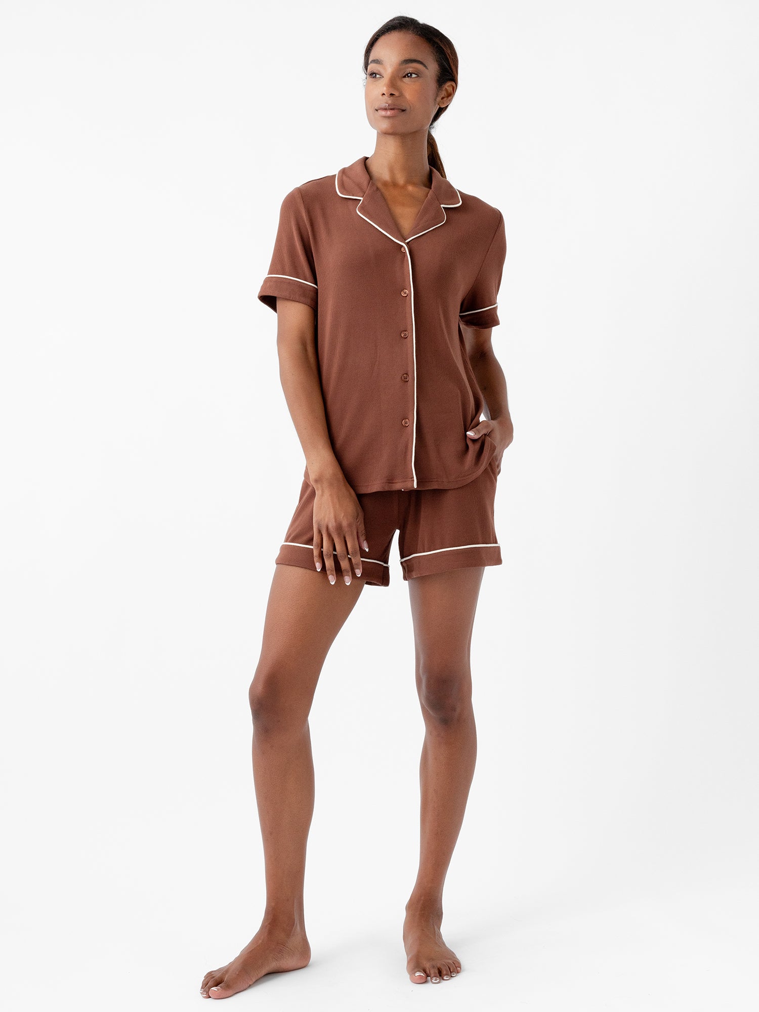 A woman is wearing Cozy Earth's Women's Bamboo Rib Knit Classic Short Sleeve Pajama Top in brown, paired with matching shorts. The pajama set features white trim along the edges. She stands barefoot against a white background, with one hand in her pocket and gazing off to the side with a relaxed expression. |Color:Spice