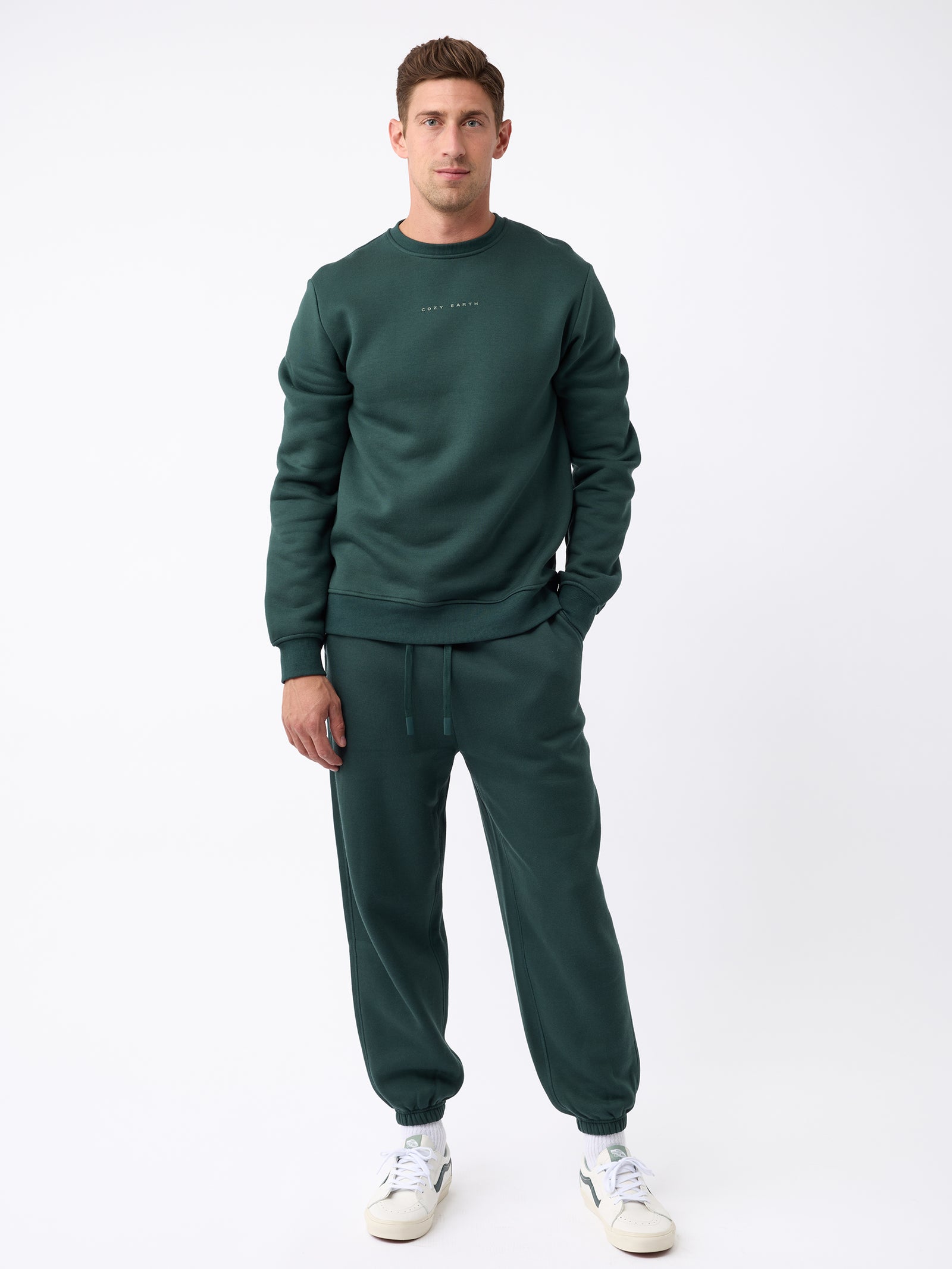 A man is standing against a white background wearing the dark green Men's CityScape Crewneck from Cozy Earth paired with matching joggers. He completes his look with white sneakers. 