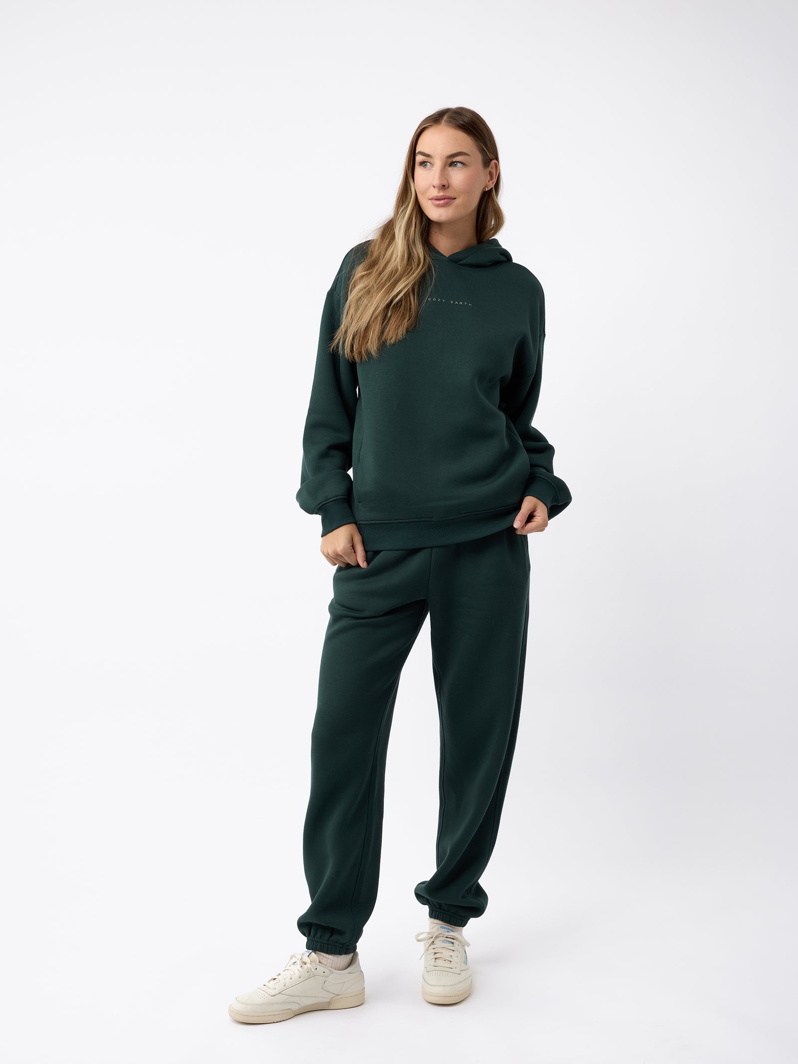 A woman stands confidently against a plain, white background, wearing a dark green CityScape Hoodie by Cozy Earth and matching jogger pants. She has long hair and is looking to the side, with her hands in her pockets and light-colored sneakers on her feet. 