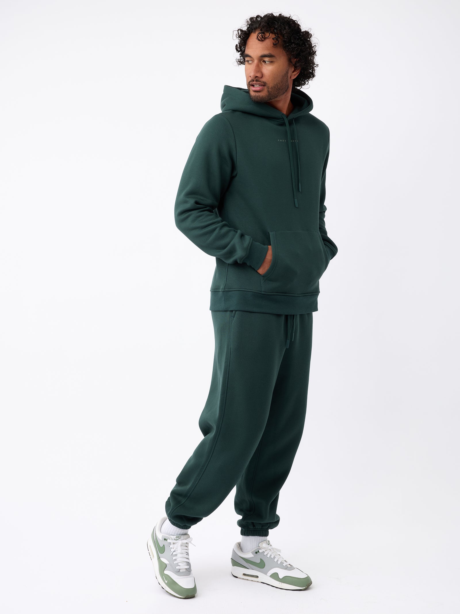 A person with curly hair wears a dark green Men's CityScape Hoodie by Cozy Earth and matching sweatpants, paired with white and green sneakers. They are set against a plain white background, looking toward the side with their hands in their hoodie pockets. 