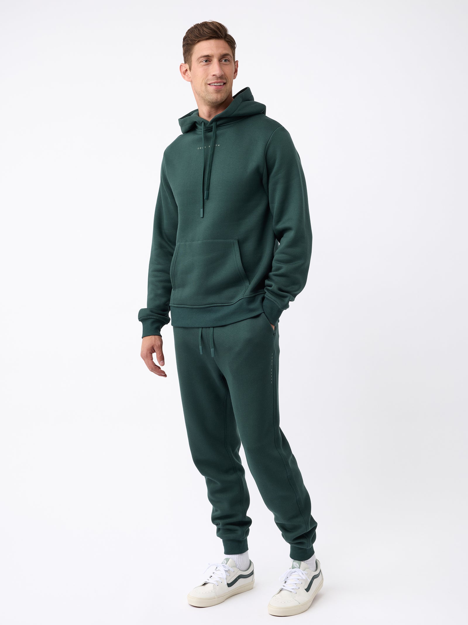 A person wearing the Men's CityScape Jogger by Cozy Earth in dark green stands against a plain white background. They are wearing white sneakers and have their hands in the jogger's hoodie pocket, looking slightly to the side. 