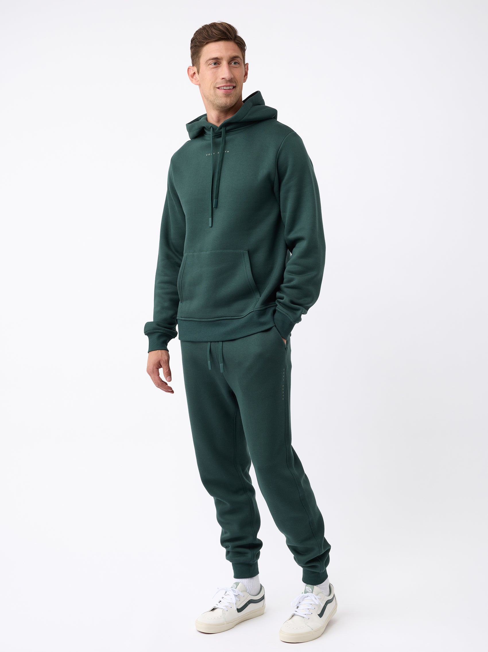 A person wearing the Men's CityScape Jogger by Cozy Earth in dark green stands against a plain white background. They are wearing white sneakers and have their hands in the jogger's hoodie pocket, looking slightly to the side. |Color:Spruce