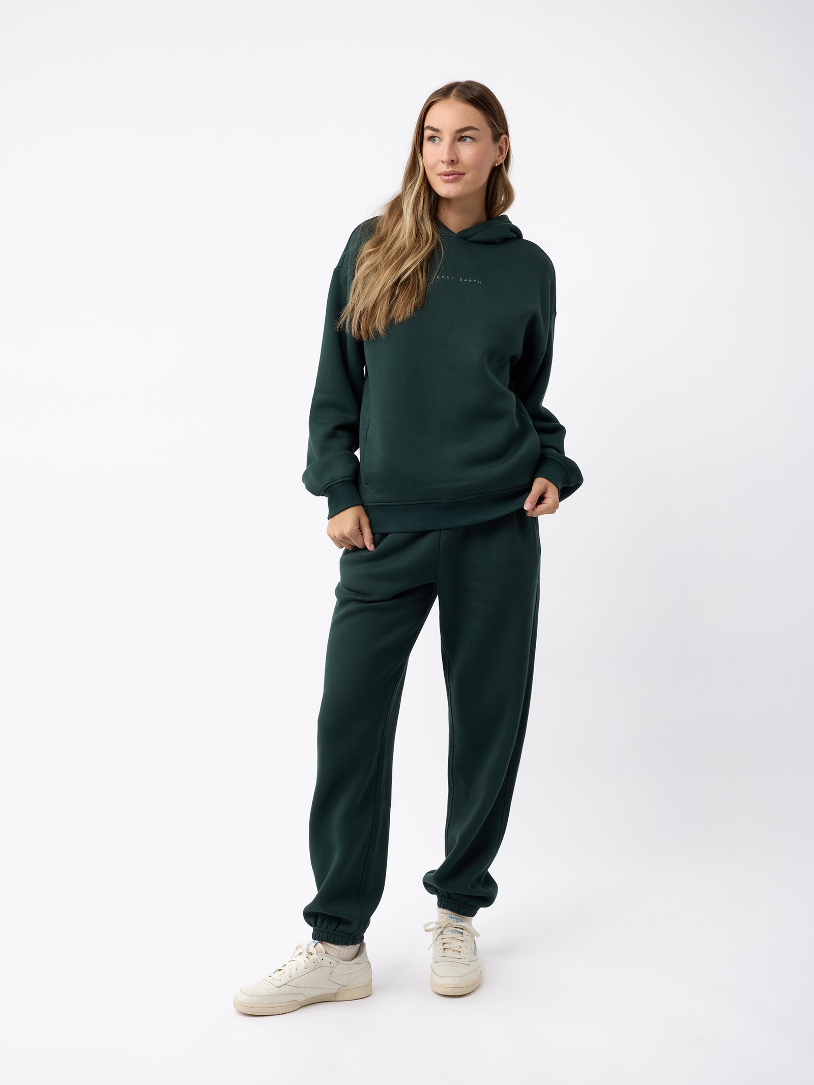 A woman stands against a plain background, wearing dark green attire from Cozy Earth, including the Women's CityScape Sweatpant. She has long, light brown hair and white sneakers. Her right hand is in her pocket as she gazes slightly to the side with a relaxed expression. 