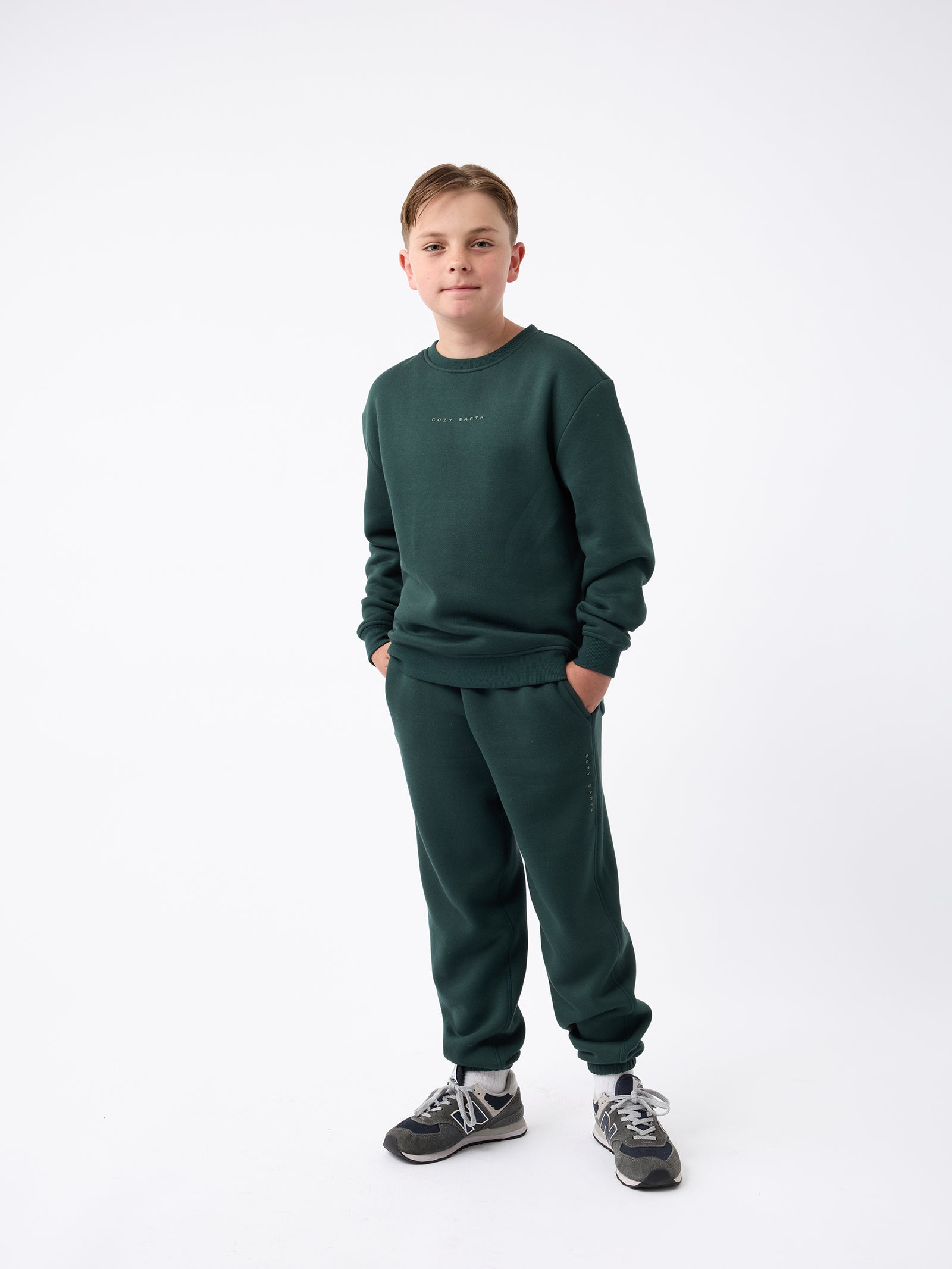 A young boy stands confidently against a white background, dressed in Cozy Earth's dark green Kid's CityScape Sweatpant with a matching sweatshirt. His hands are in his pockets as he looks directly at the camera, complementing his outfit with gray sneakers. 