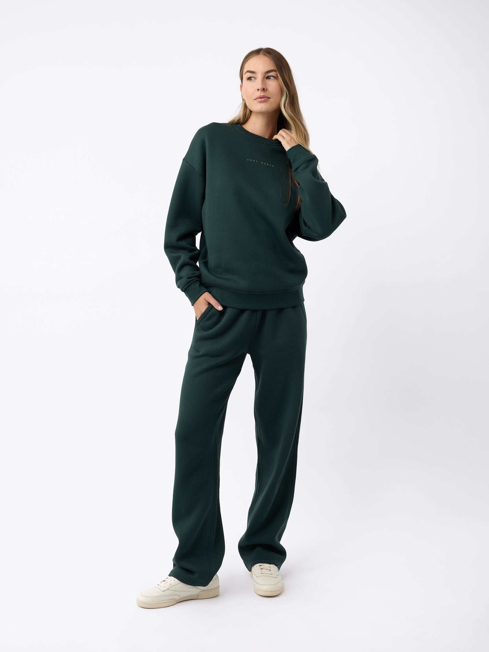 A person wearing the Women's CityScape Wide Leg Pant by Cozy Earth in a dark green color stands against a plain white background. They have long hair and are wearing white sneakers, with one hand in their pocket and a relaxed pose. 