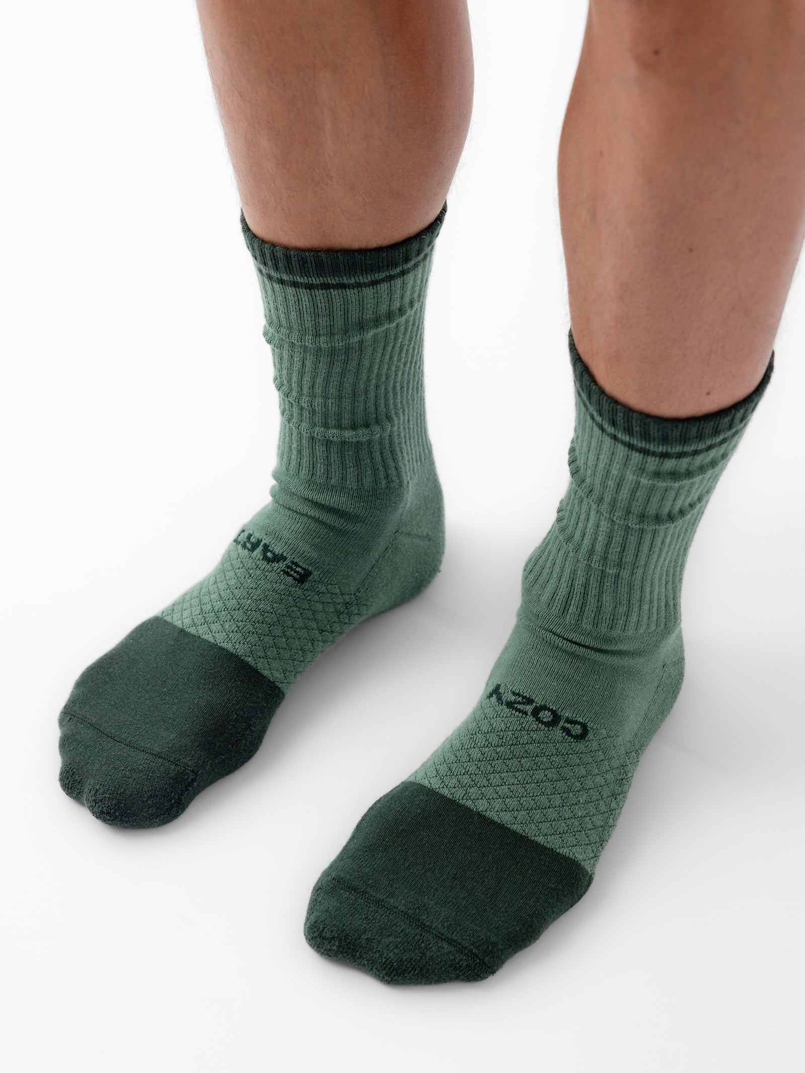 Close-up of a person wearing a pair of green Essential Calf Socks by Cozy Earth. The socks, shown on feet standing on a white surface, feature different shades of green with ribbed and elastic material around the ankle area. 