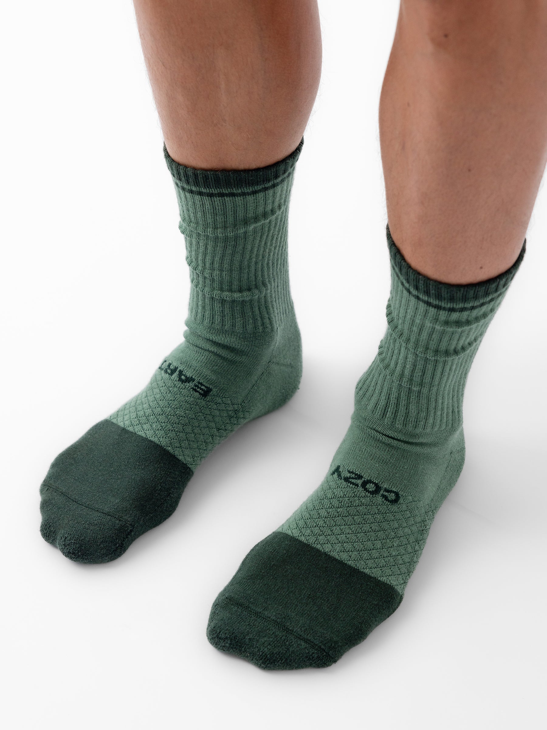 Close-up of a person wearing a pair of green Essential Calf Socks by Cozy Earth. The socks, shown on feet standing on a white surface, feature different shades of green with ribbed and elastic material around the ankle area. |Color:Spruce