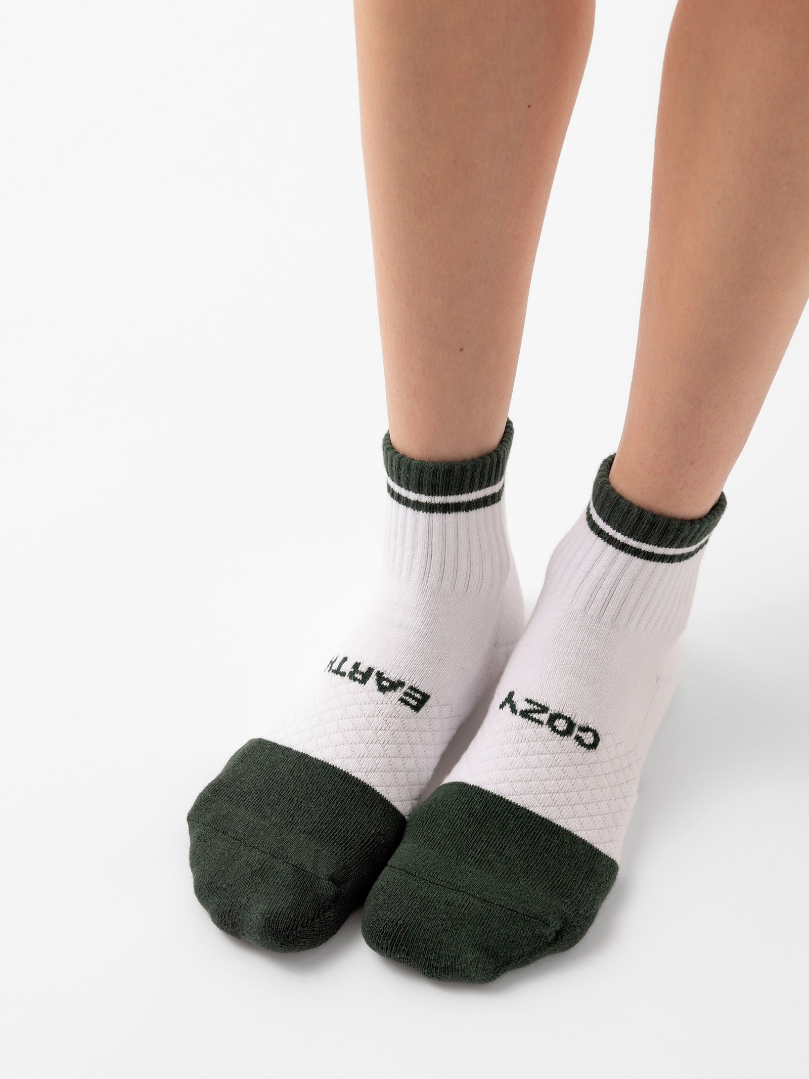 Two legs wearing socks from the Cozy Earth Essential Quarter Sock 2-Pack collection. The white ankle socks feature dark green toes and cuffs, with "EARTH" written in black on the left foot and "COZY" on the right foot against a plain white background. 