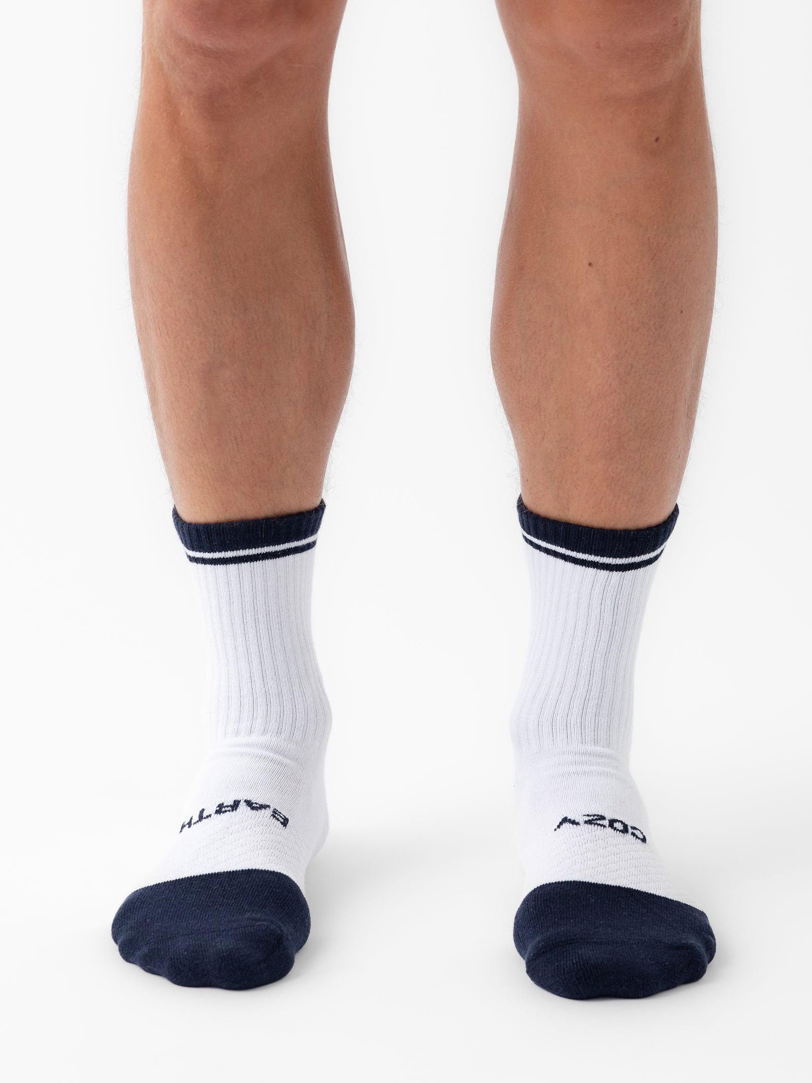 Close-up of a person's lower legs wearing white athletic socks with dark blue cuffs and toes from the Essential Calf Sock 4-Pack by Cozy Earth. The left sock has "EARTH" written on the toes, and the right sock has "AZOS" written on the toes. The background is plain white. 