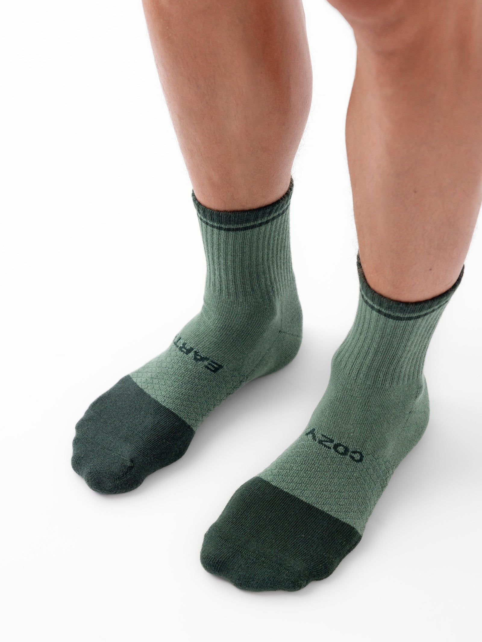 A person wearing green quarter socks from the Cozy Earth Essential Quarter Sock 4-Pack, featuring darker green accents at the toes, heels, and cuffs. The socks are adorned with the words "EARTH" and "COZY" near the toes. The person's lower legs are visible against a white background. 