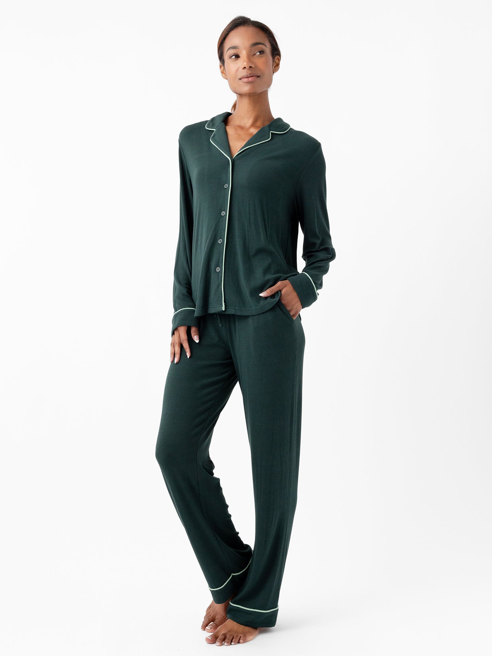 A person stands barefoot against a plain white background, sporting Cozy Earth's Women's Bamboo Rib Knit Classic Pajama Pant in matching dark green with white piping trim. The set comprises a long-sleeve button-up top with a collar and long pants, with the person's hands nestled in the pockets of the pants. 