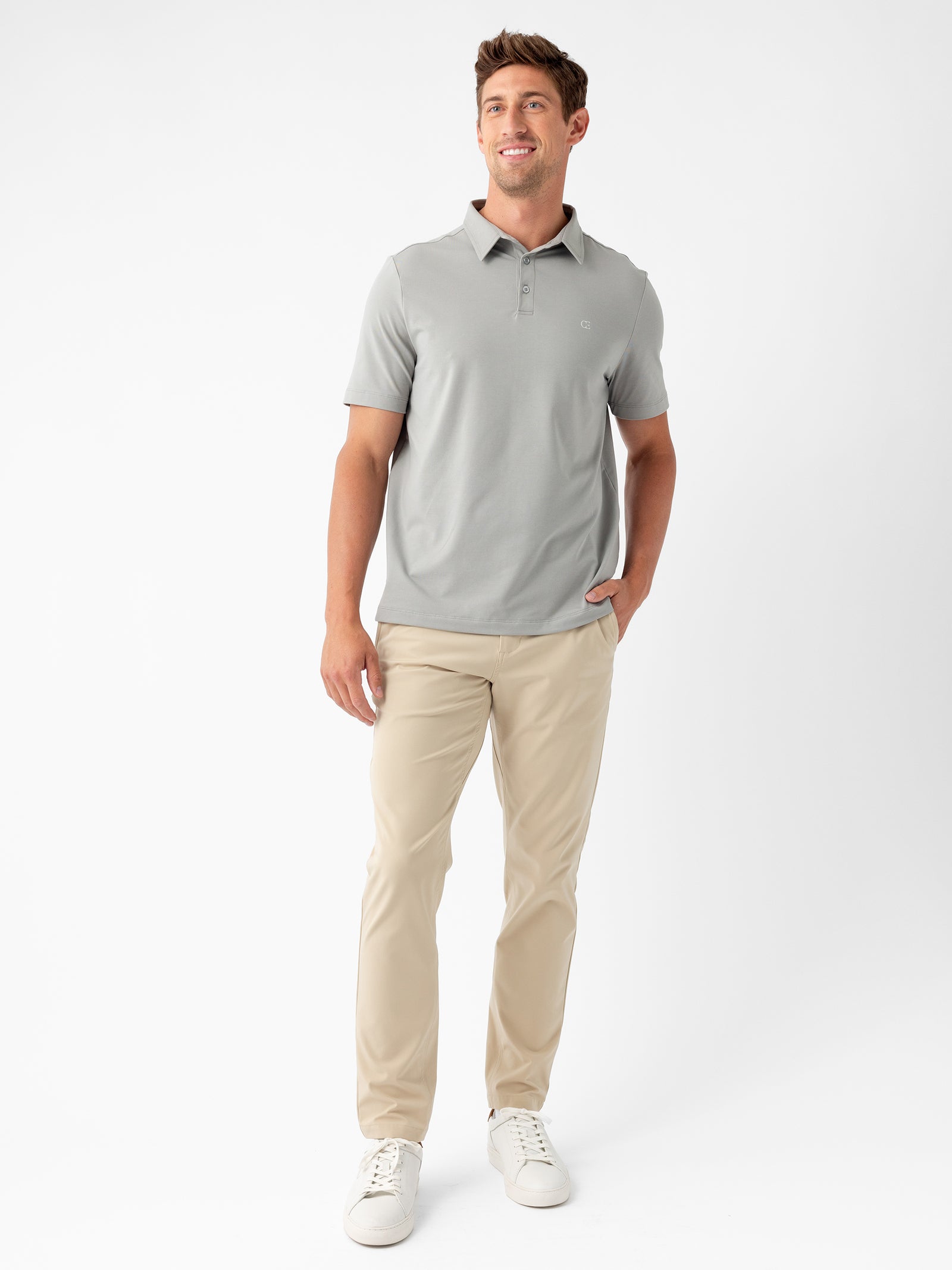 A man with short brown hair stands against a white background, smiling as he wears Cozy Earth's Men's Everyday Polo in light gray paired with beige pants. His left hand rests in his pocket, and he completes his outfit with white sneakers. 