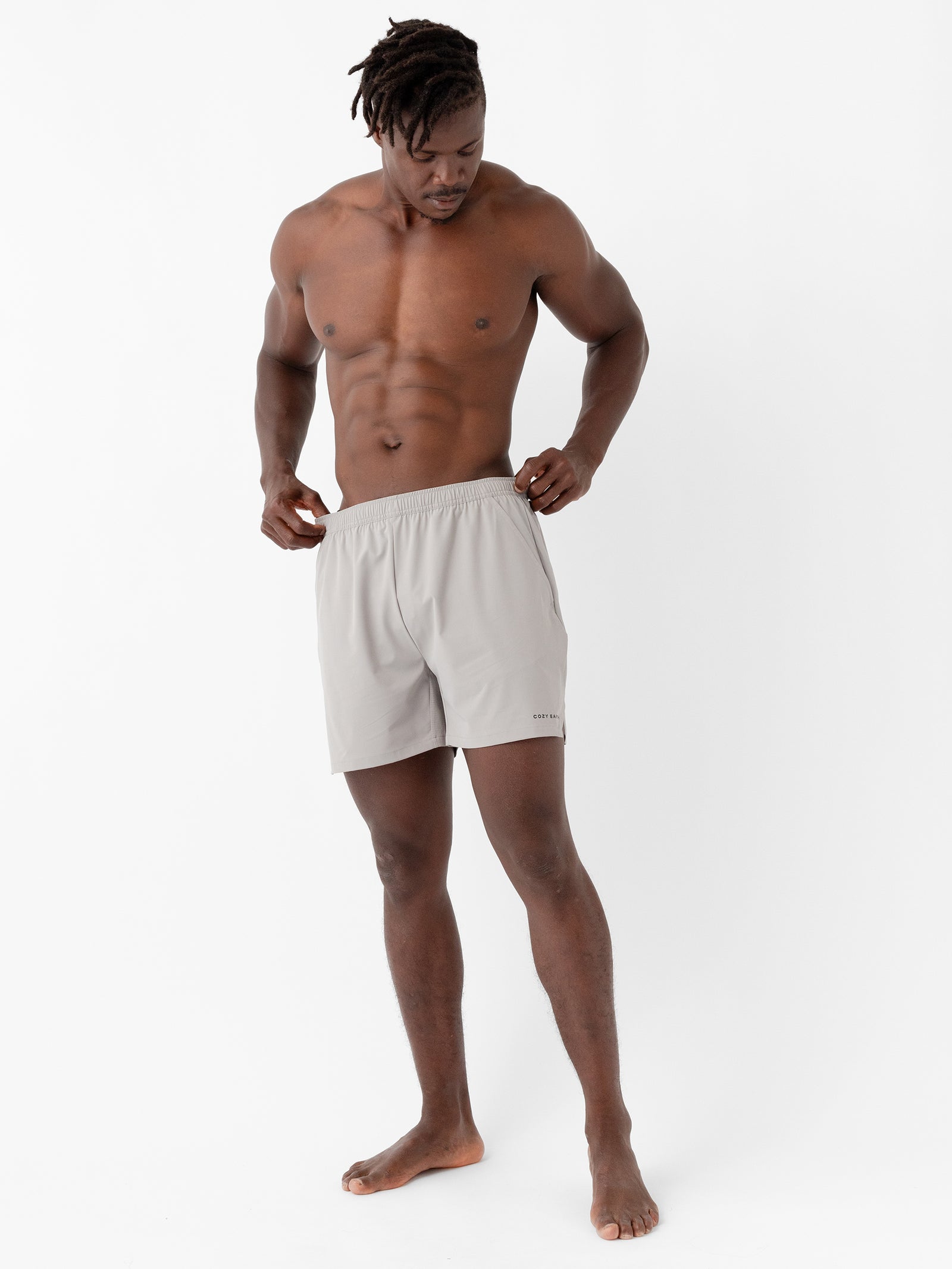 A man with a muscular build and dreadlocks stands against a plain white background. He is wearing light gray Men's Performance Sleep Shorts by Cozy Earth and looking down while slightly adjusting them with his hands. 