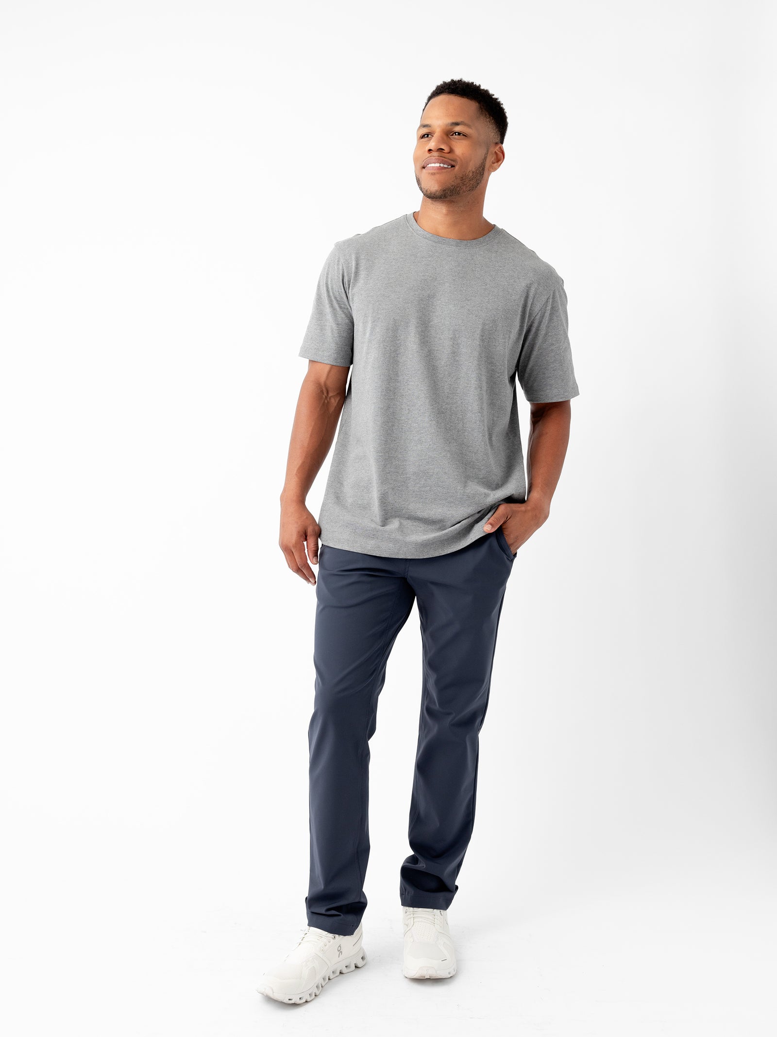 A man is standing against a plain white background, wearing the Men's All Day Tee by Cozy Earth in gray, paired with dark blue pants and white sneakers. He has a slight smile and his right hand is in his pocket while his left hand hangs by his side. 