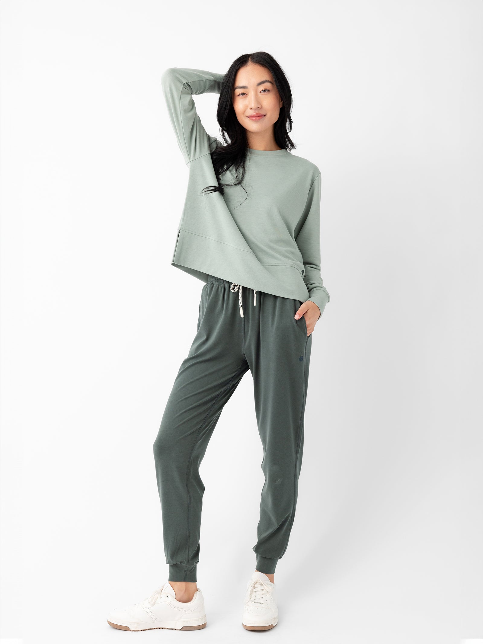 A woman stands against a plain white background wearing Cozy Earth’s Women's Studio Jogger set in light green. She smiles softly with one hand on her hip and the other behind her head, complementing her outfit with white sneakers. 