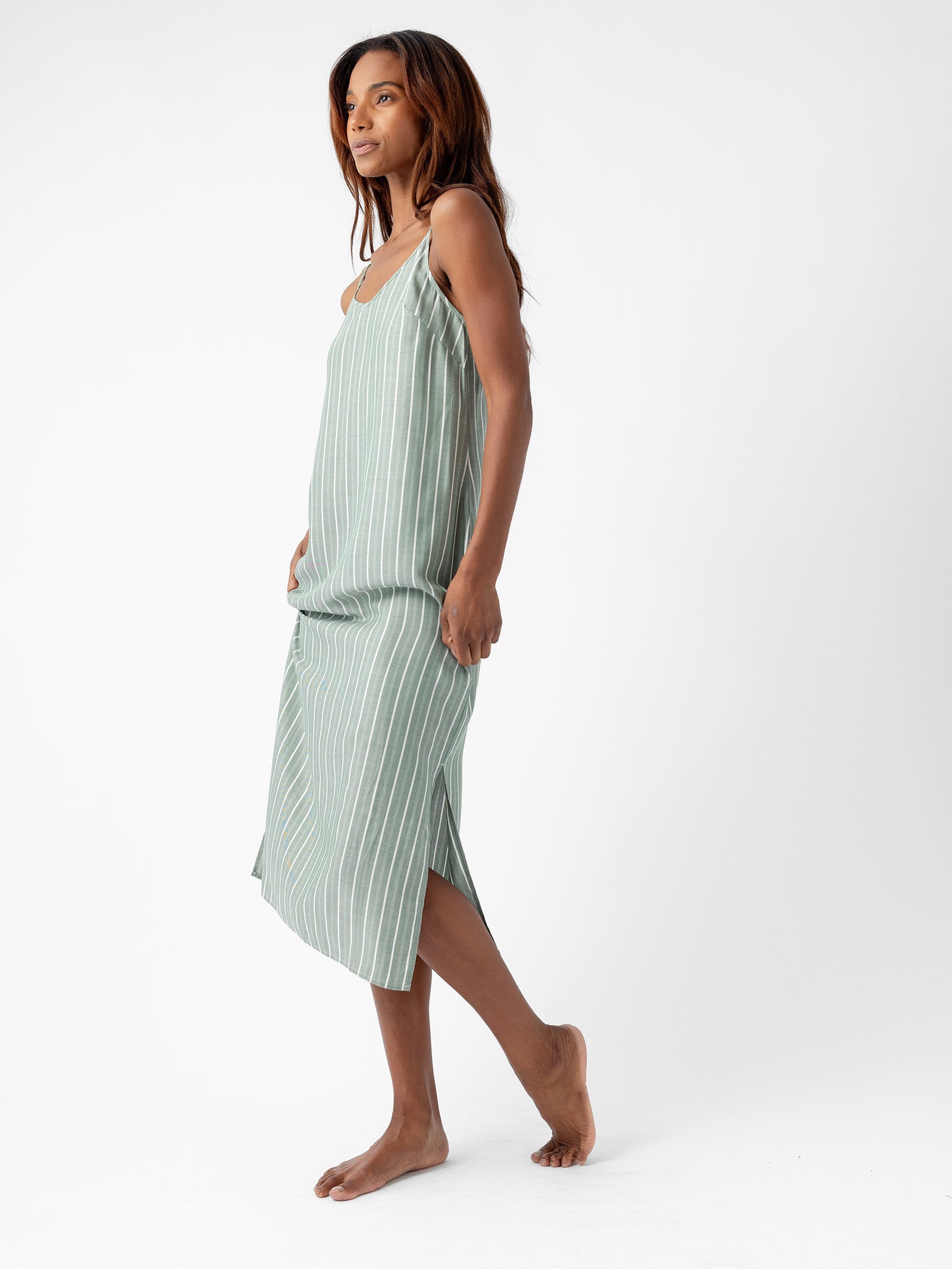 A woman stands barefoot on a plain white background, wearing the Women's Soft Woven Nightgown from Cozy Earth. The nightgown is green with white vertical stripes and features a side slit. She gazes to her left with a relaxed expression. 