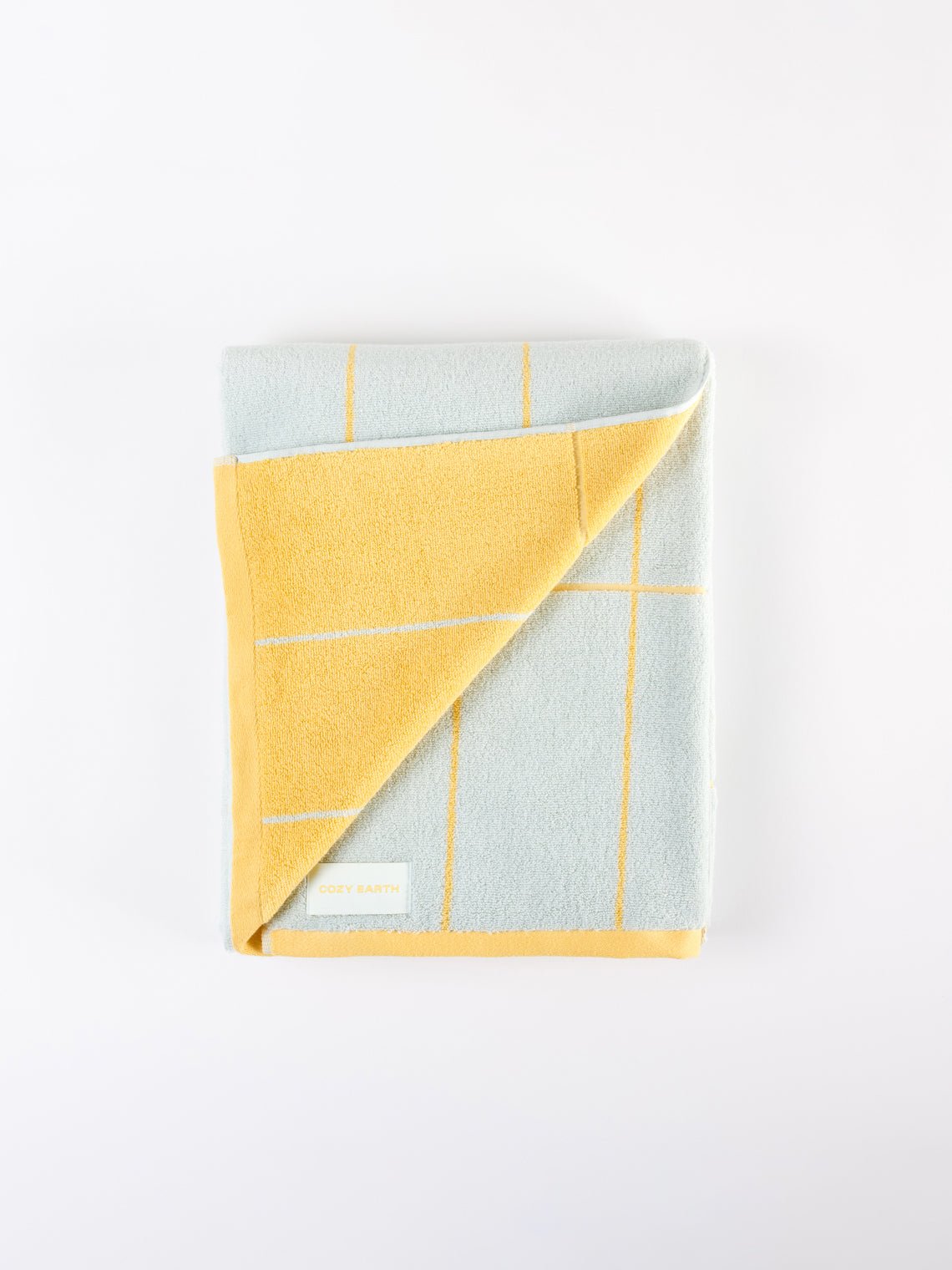 The Windowpane Resort Towel by Cozy Earth is displayed folded, showcasing a light gray and yellow checkered design. One corner is flipped to reveal the yellow underside, and a label with text is near the corner on a white background. |Color:Sunny Sky