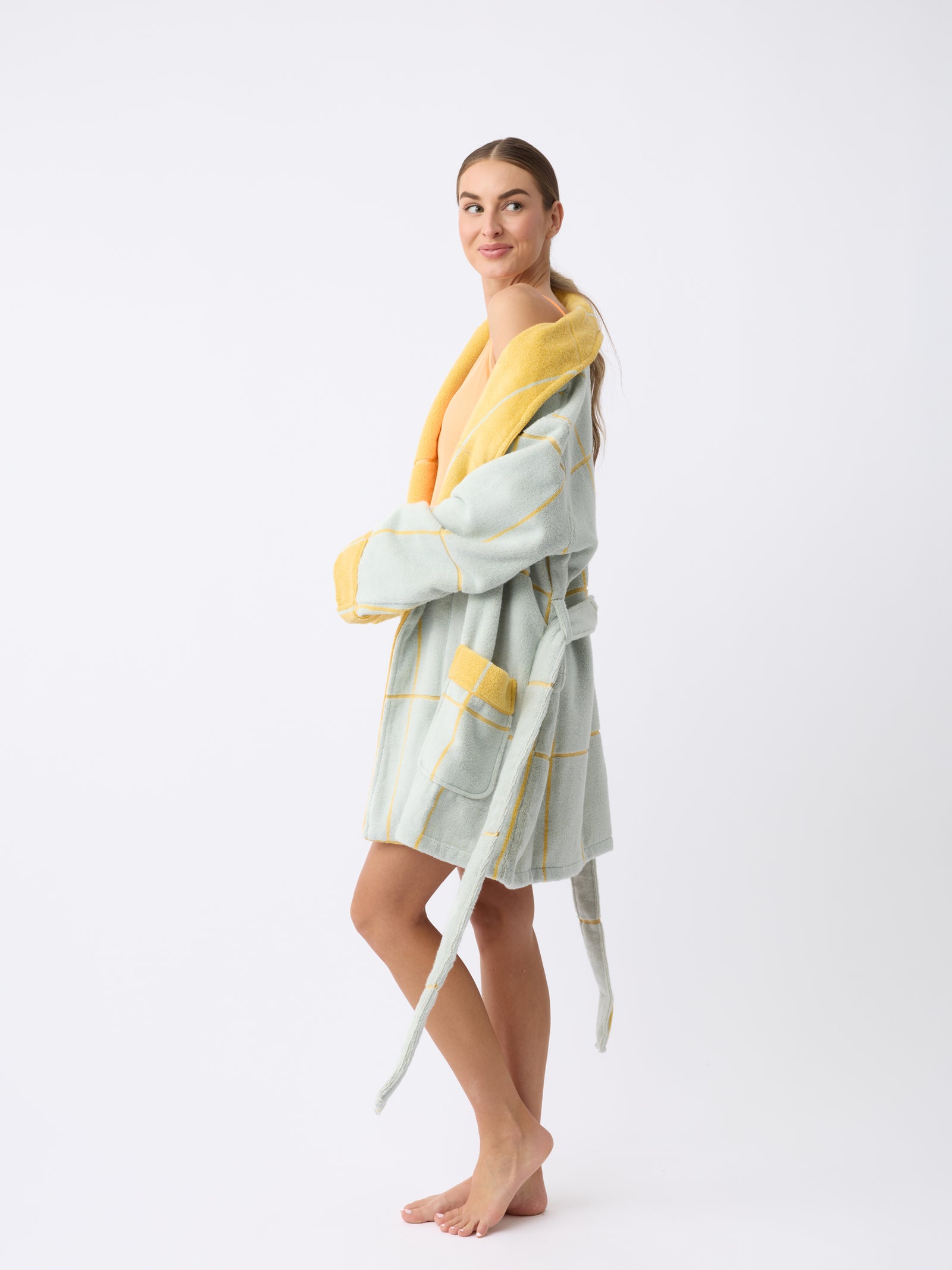 A woman smiles, wearing Cozy Earth's Windowpane Resort Robe in light blue with yellow trim and large pockets. She stands barefoot on a plain white background, the robe's belt loosely tied around her waist. 