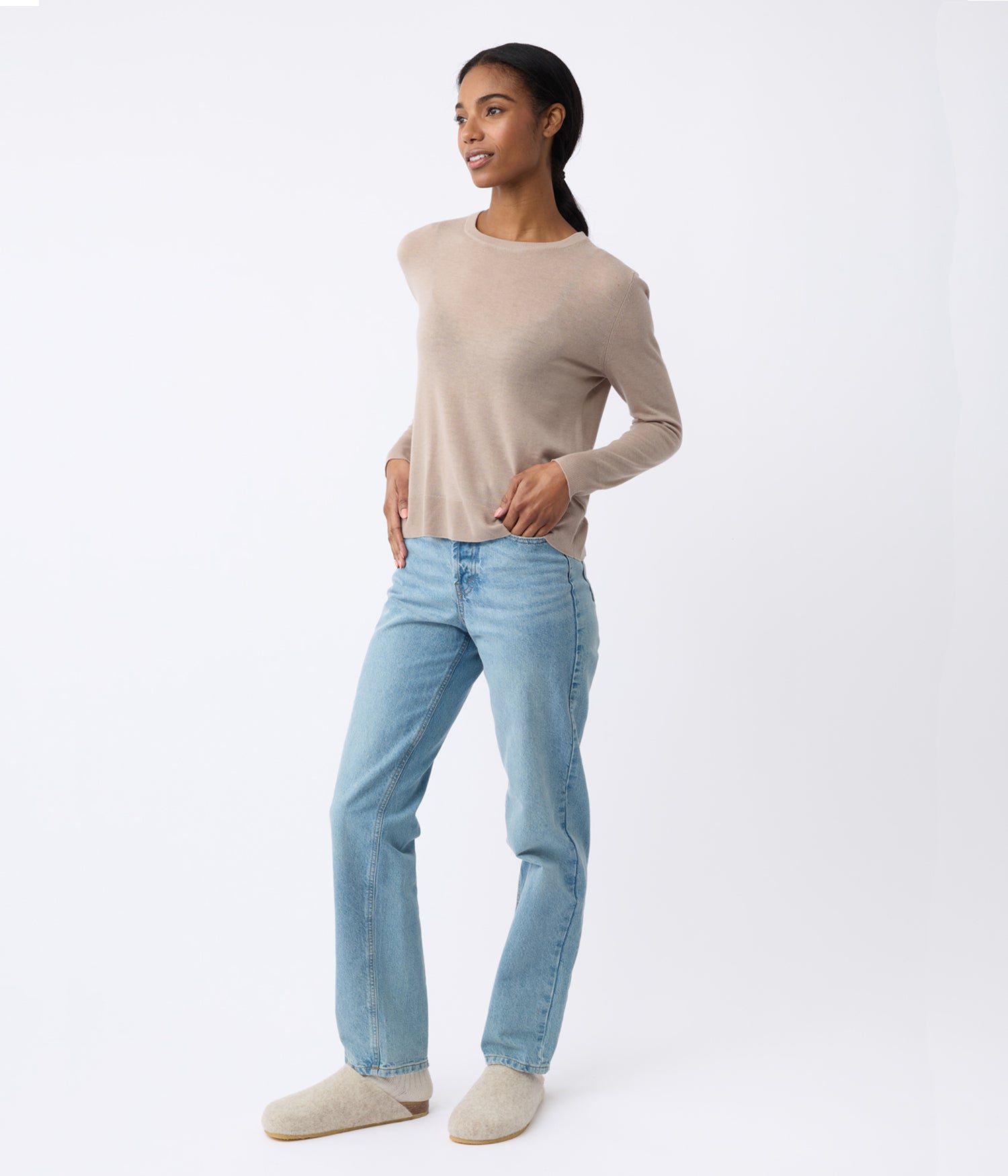 The woman, dressed in a Cozy Earth's Women's AirKnit Crewneck Sweater, light blue jeans, and cream-colored slip-ons, stands against a white background. With their hands in pockets and facing slightly to the side, they exhibit a relaxed expression. |Color:Taupe