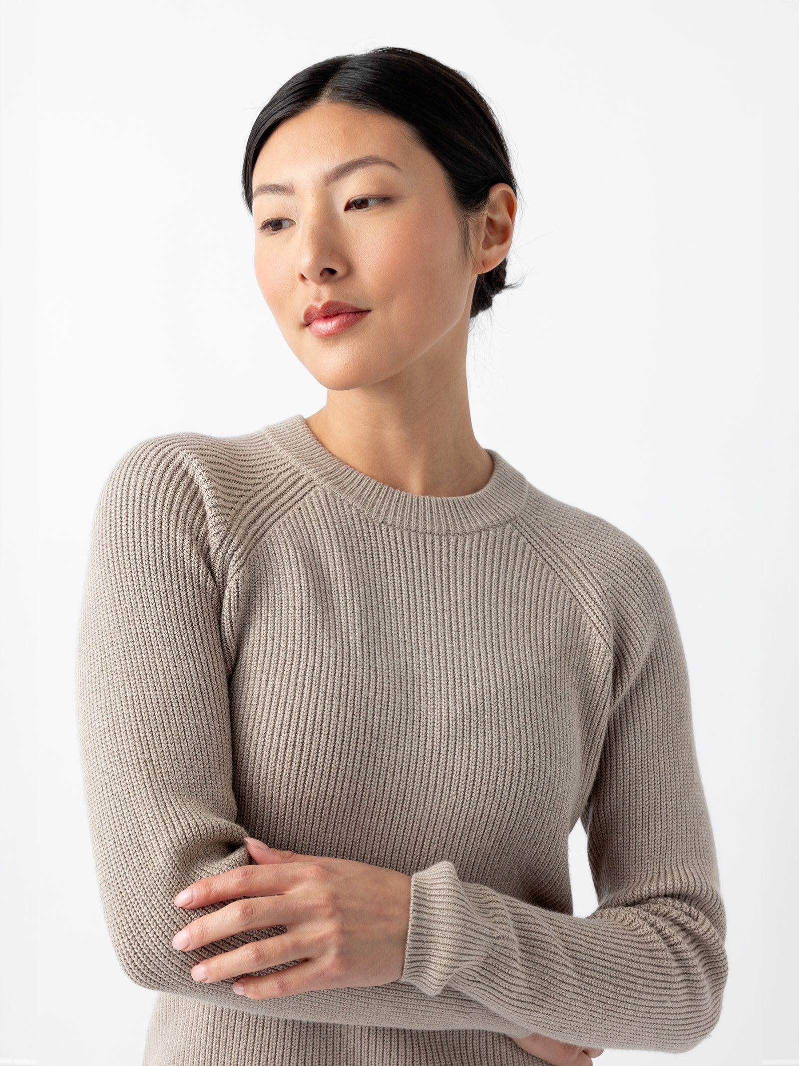 A person with dark hair pulled back is wearing a light gray, long-sleeved Women's Classic Crewneck from Cozy Earth. They are looking to the side with a neutral expression and have their arms crossed gently in front of them. The background is plain white. 