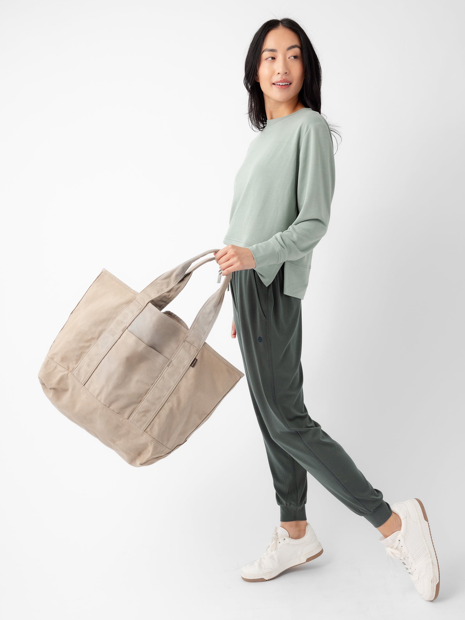 A woman with long dark hair is posing against a plain white background. She wears a light green long-sleeve top, dark green joggers, and white sneakers while carrying the Waxed Canvas Tote from Cozy Earth in beige. With a slight smile, she looks to the left, exuding a relaxed and casual demeanor. 