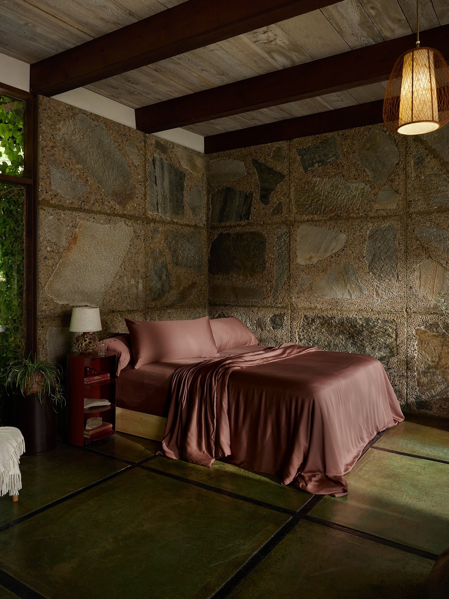 A cozy bedroom with a stone wall, featuring a bed adorned with Cozy Earth's Bamboo Duvet Cover in pink, surrounded by matching pillows. The wooden ceiling boasts exposed beams, and a wicker lamp hangs elegantly. A small table stands beside the bed, complete with a lamp and potted plant, while the floor showcases greenish-tinted concrete. 