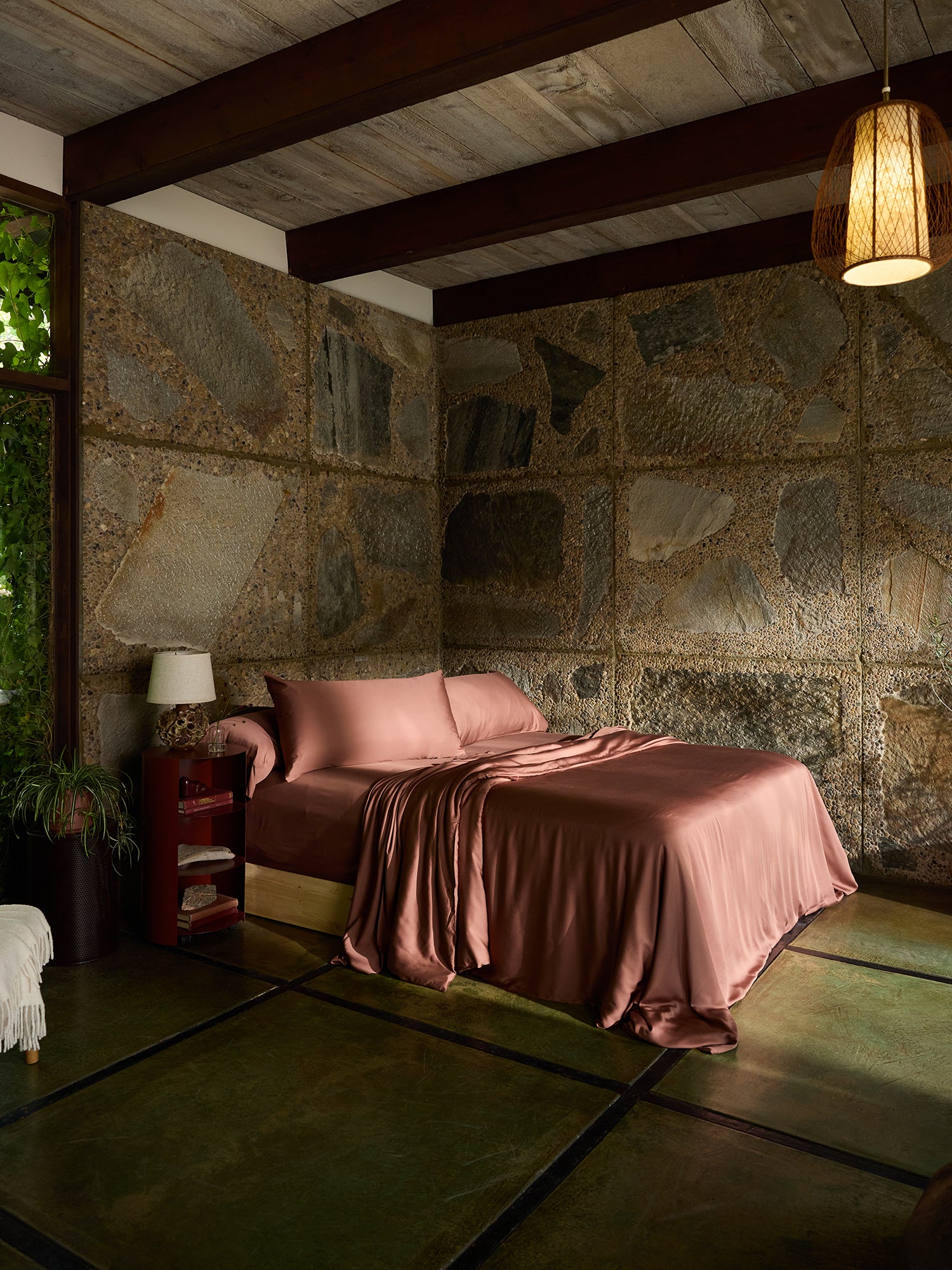 A cozy bedroom with rustic stone walls, a wooden ceiling with exposed beams, and a green-tinted concrete floor. The bed is topped with an elegant set of mauve-colored Bamboo Fitted Sheets by Cozy Earth and a matching duvet. A red side table with a white lamp and potted plants adorn the corner. 