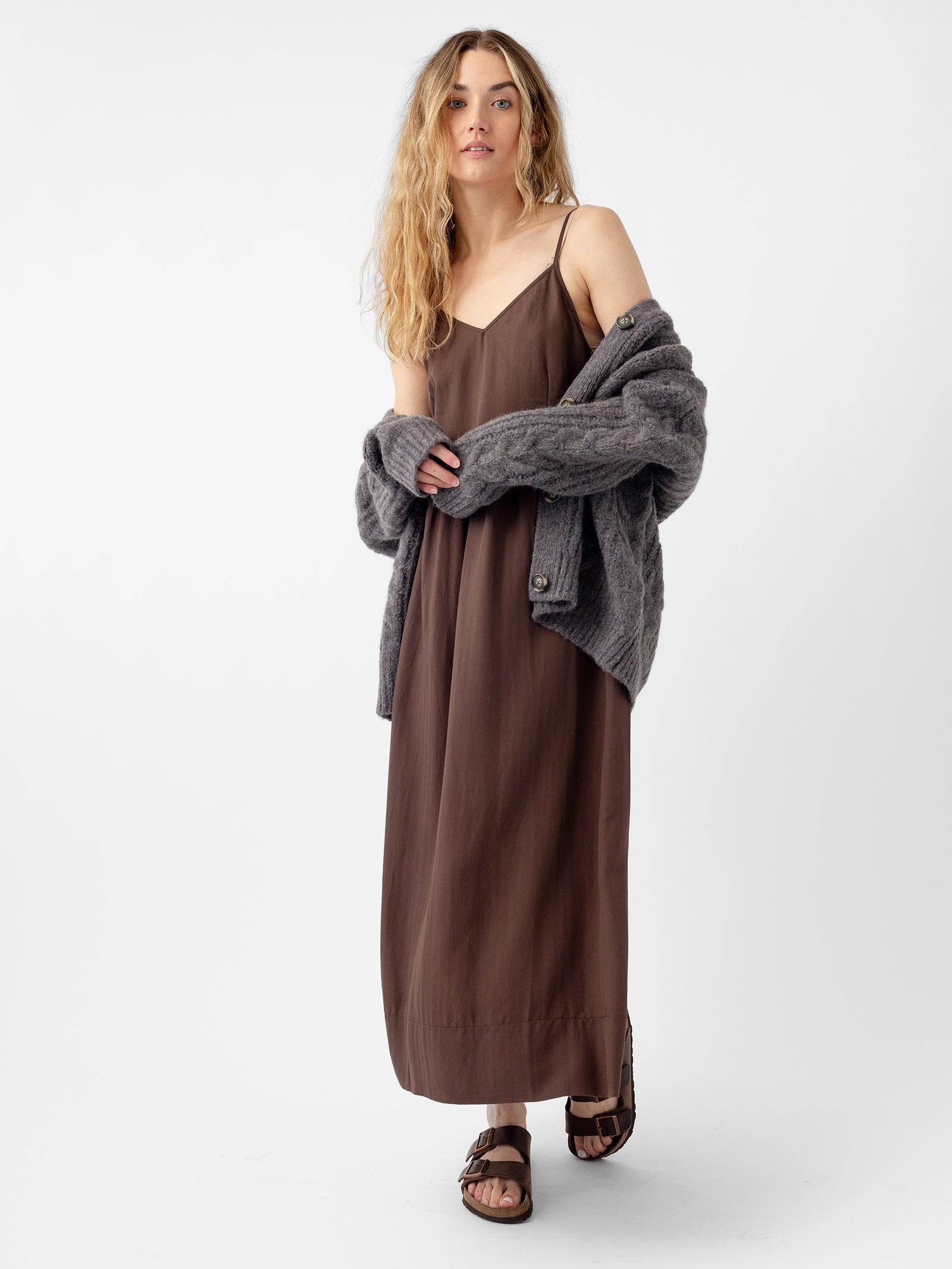 A person with long blond hair is wearing a brown sleeveless dress paired with the Oversized Cable Knit Cardigan from Cozy Earth and sandals, standing against a plain white background. 