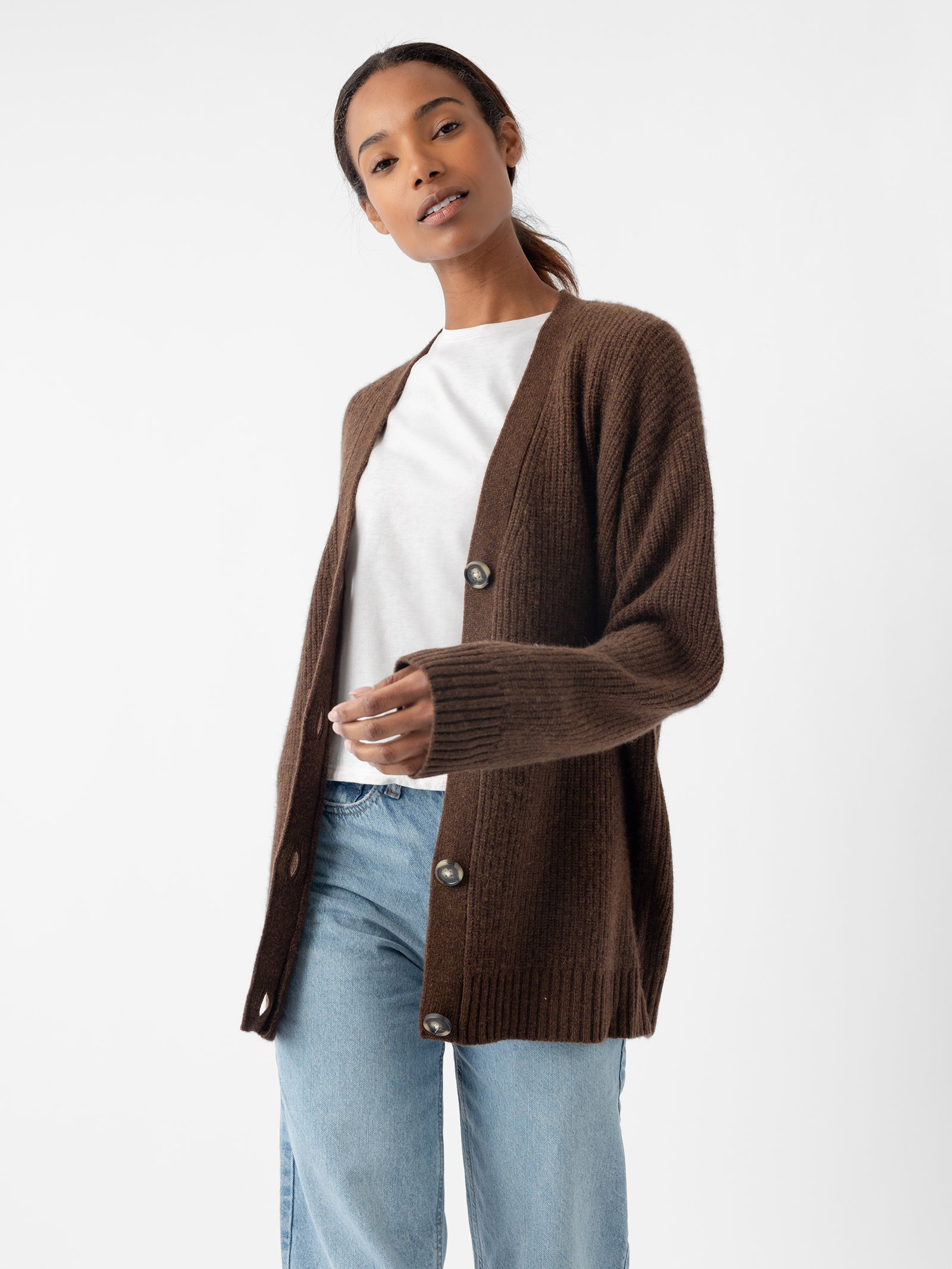 A model showcases the Oversized Sunday Cashmere Cardigan by Cozy Earth, a brown buttoned cardigan, while wearing a white shirt and blue jeans against a plain white background. 