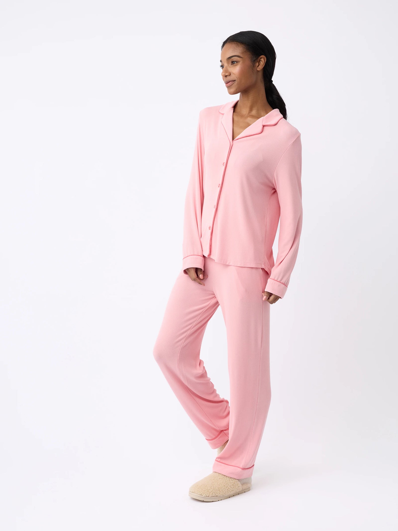 A woman is wearing Cozy Earth's pink Women's Bamboo Rib-Knit Classic Pajama Pant set with slippers, standing against a plain white background. The pajamas feature a button-up top with a collar and matching pants, making the individual appear relaxed and comfortable. 