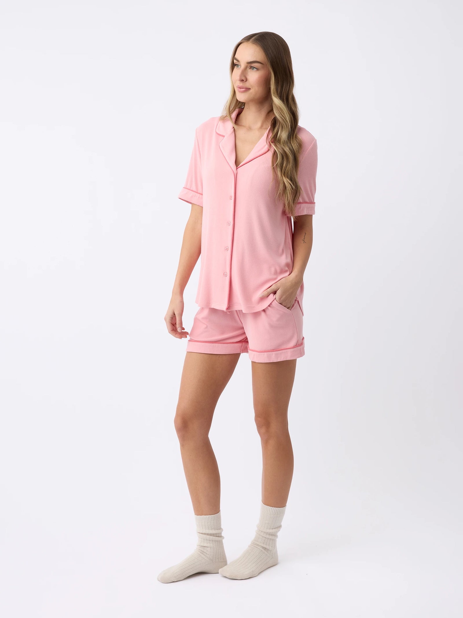 A woman in a Cozy Earth Women's Rib-Knit Classic Short Sleeve Pajama Set, featuring a pink button-up top and matching shorts, stands against a plain white background. She has long wavy hair, looks to the side, and wears white socks. 