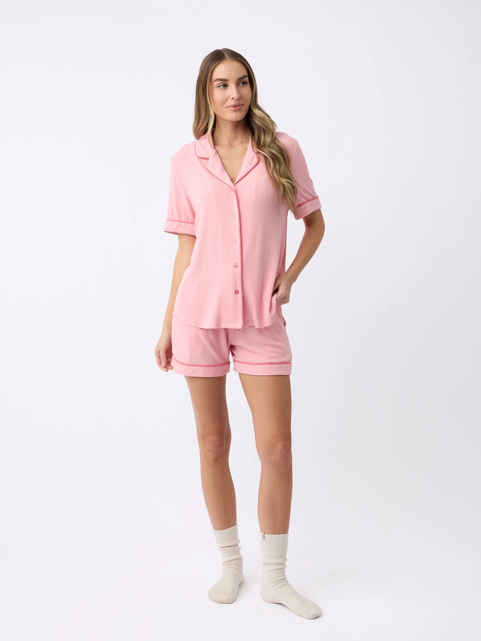 A woman in Cozy Earth's pink Bamboo Rib-Knit Classic Short Sleeve Pajama Top and matching shorts poses casually with one hand in her pocket against a white background, wearing light-colored socks. 