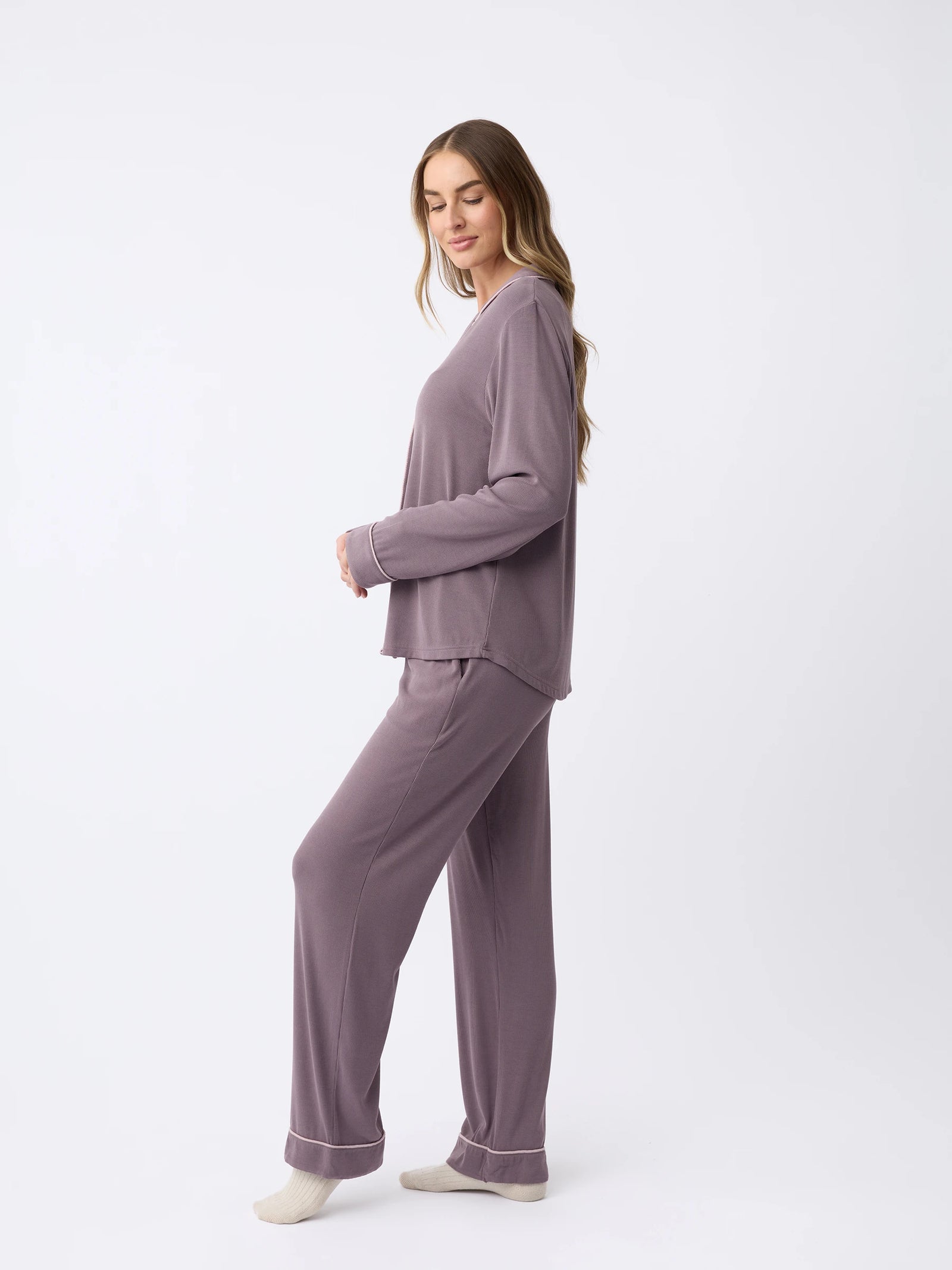 A woman with long hair stands sideways wearing the Rib-Knit Classic Long Sleeve Pajama Set by Cozy Earth. The matching gray outfit includes a long-sleeved top and pants. She looks down with a neutral expression against a plain white background. 