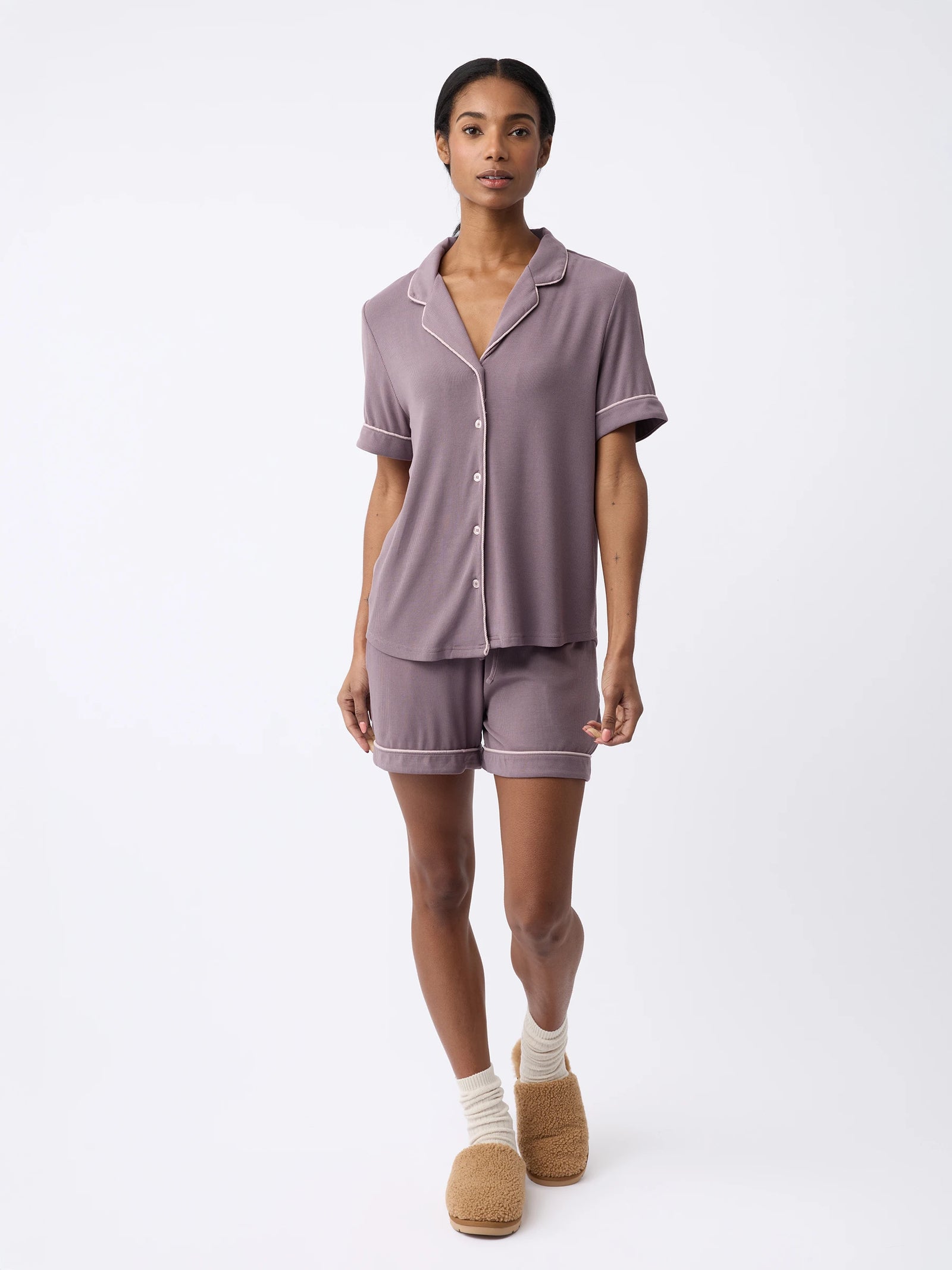 A woman wearing Cozy Earth's Women's Bamboo Rib-Knit Classic Pajama Short set in muted purple, featuring white piping. They're also in light-colored socks and fuzzy tan slippers, standing against a plain white background. 