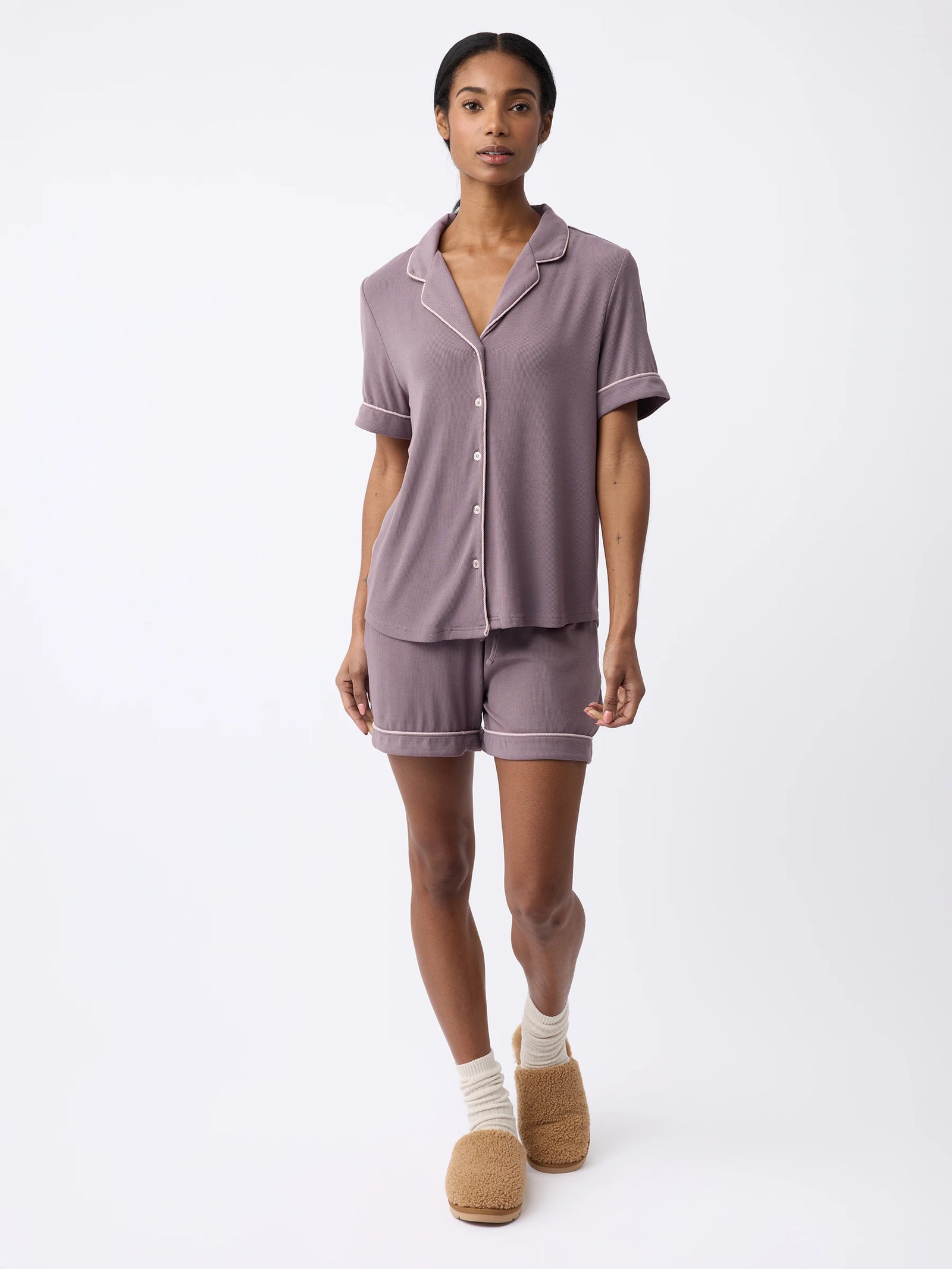 A woman is dressed in a Cozy Earth Women's Bamboo Rib-Knit Classic Short Sleeve Pajama Top and matching shorts, paired with beige socks and tan slippers, standing against a white background. 