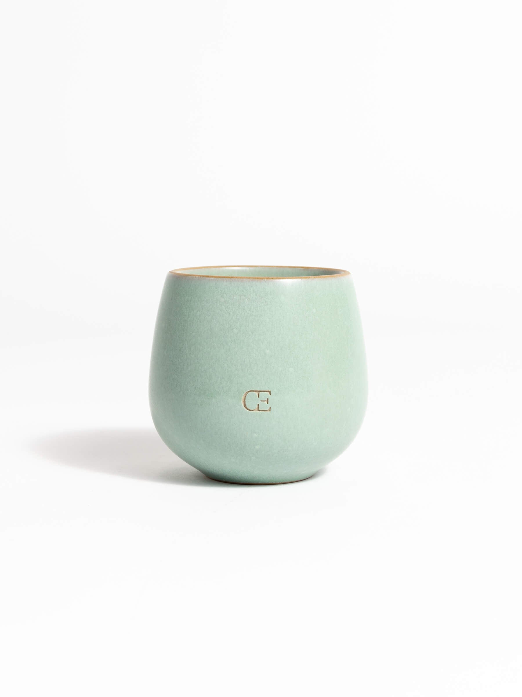 A light green ceramic Candle UNWIND by Cozy Earth, featuring a rounded shape and a small subtle logo on the front, set against a plain white background.