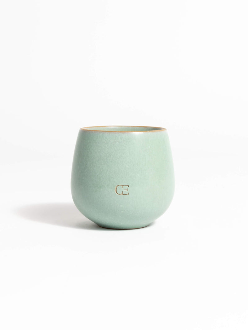 A light green ceramic Candle UNWIND by Cozy Earth, featuring a rounded shape and a small subtle logo on the front, set against a plain white background.