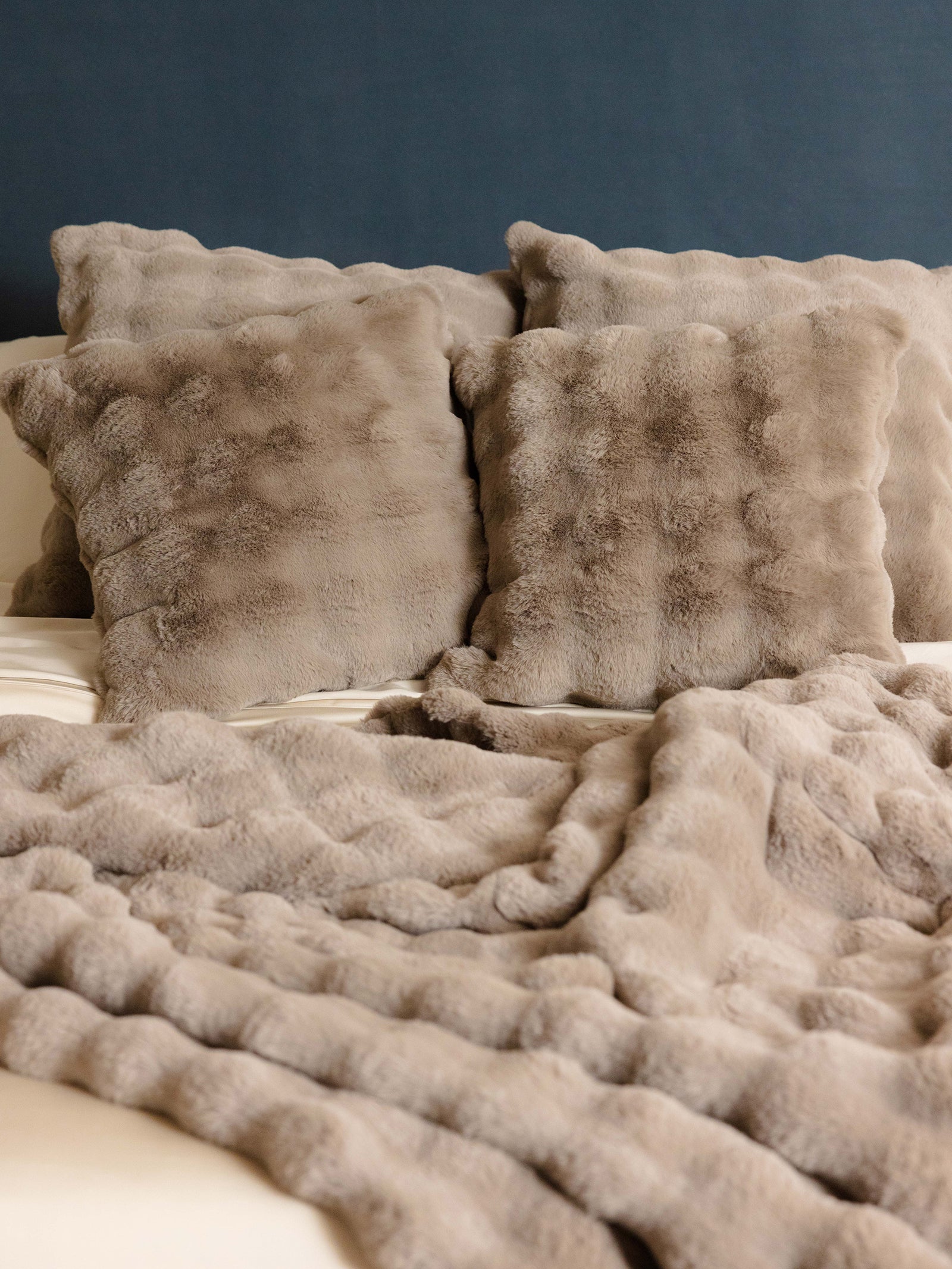 A cozy bed features the Bubble Cuddle Blanket by Cozy Earth, with multiple soft, fluffy beige pillows and a matching blanket draped over white sheets, set against a dark blue headboard. 