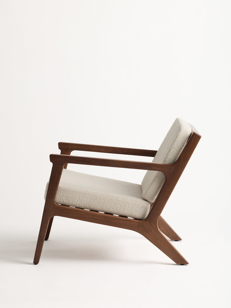 A side view of the Coronado Lounge Chair by Cozy Earth showcases its modern wooden design, featuring a light beige cushioned seat and backrest. The chair boasts sleek lines and angled armrests, all set against a plain white background.