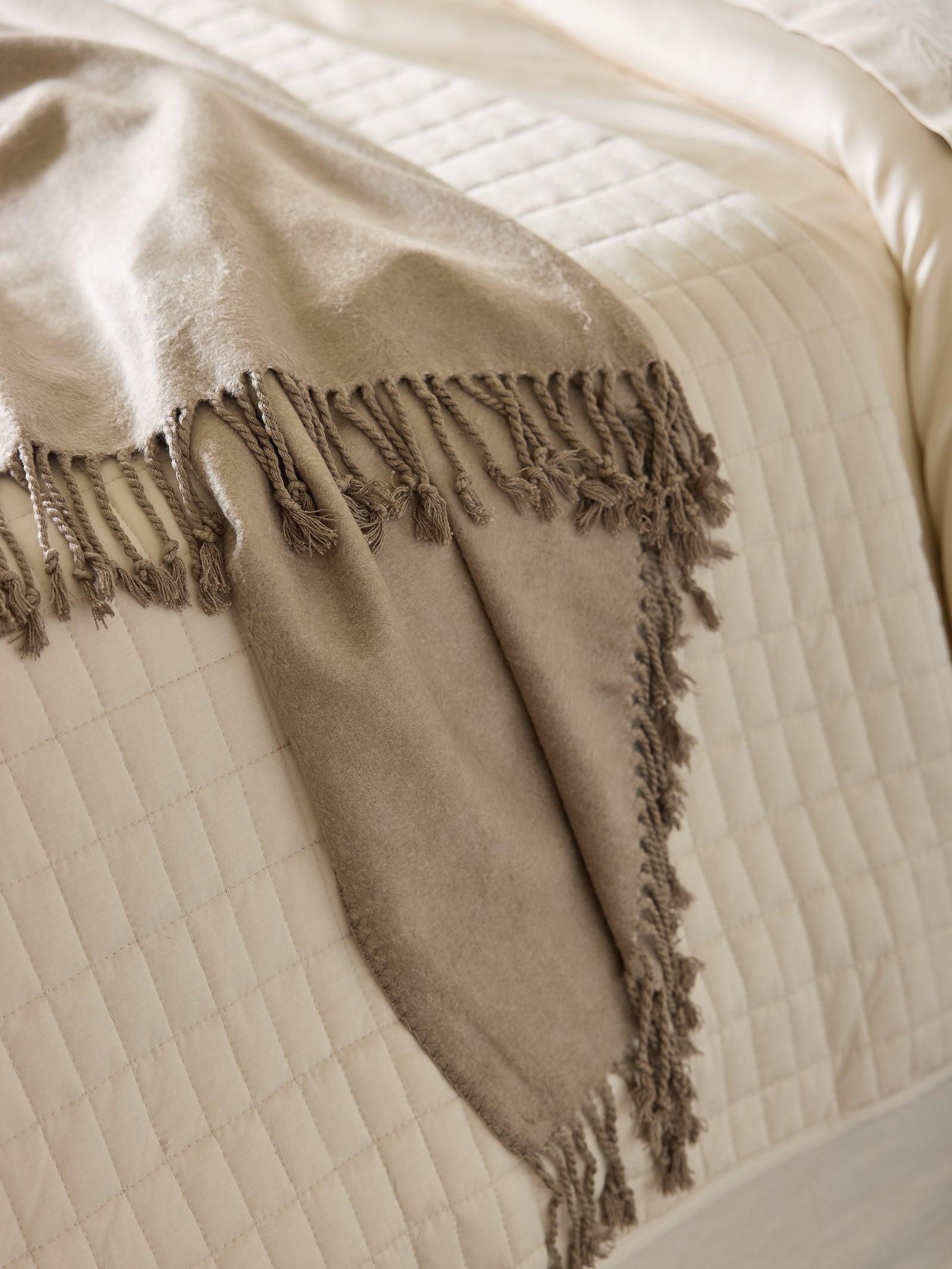 A Beige Cozy Earth Bamboo Tassel Throw is draped over the corner of a neatly made bed with a cream-colored, quilted bedspread. The fabric of the throw appears soft and cozy, contrasting subtly with the smooth texture of the bedspread. 