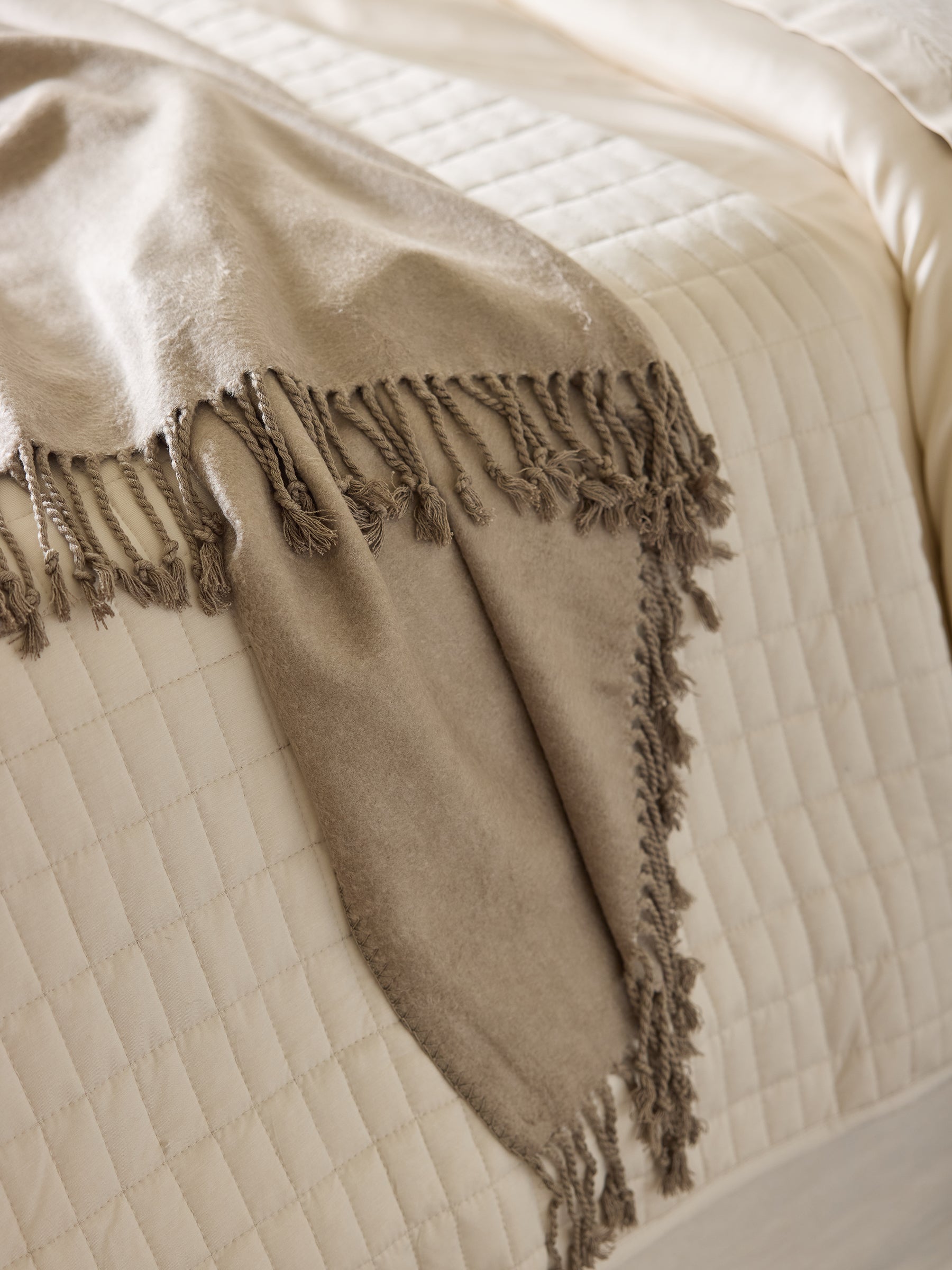 A Beige Cozy Earth Bamboo Tassel Throw is draped over the corner of a neatly made bed with a cream-colored, quilted bedspread. The fabric of the throw appears soft and cozy, contrasting subtly with the smooth texture of the bedspread. |Color:Walnut