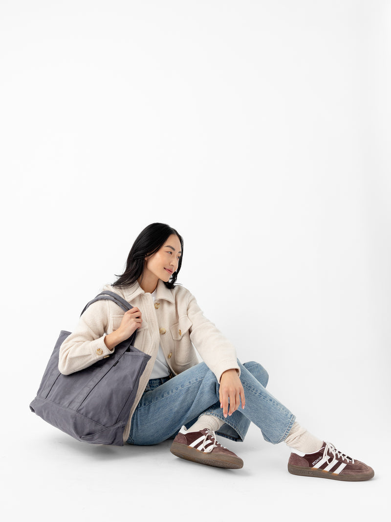 A woman dressed in a cream-colored sweater and blue jeans sits on the ground with her legs crossed. She holds a large Waxed Canvas Tote from Cozy Earth and is wearing brown sneakers. The background is a plain white studio setting.